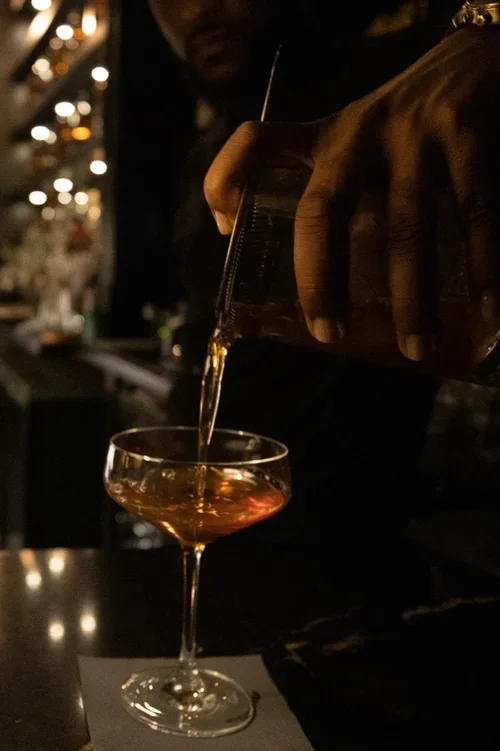 A person pouring a drink into a cocktail glass at a bar, with blurred lights in the background.