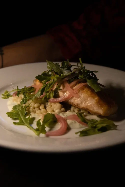A plated dish featuring a piece of grilled fish topped with mixed greens and pink onions, served with a side of small pearl-like grains, possibly couscous or quinoa.