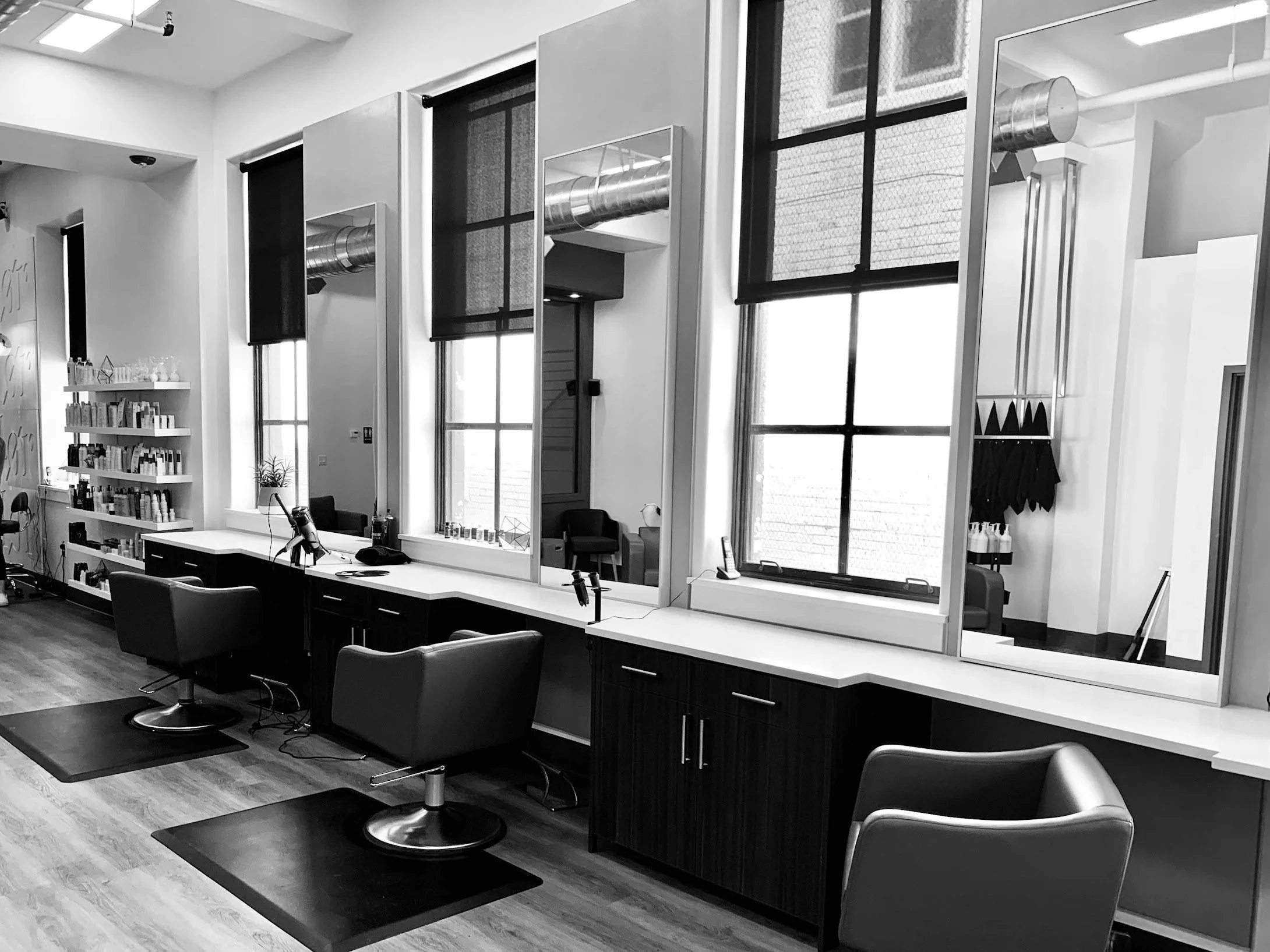 Interior of a modern hair salon or barbershop with hair styling stations, chairs, mirrors, and shelves with hair products. Large windows with black shades.