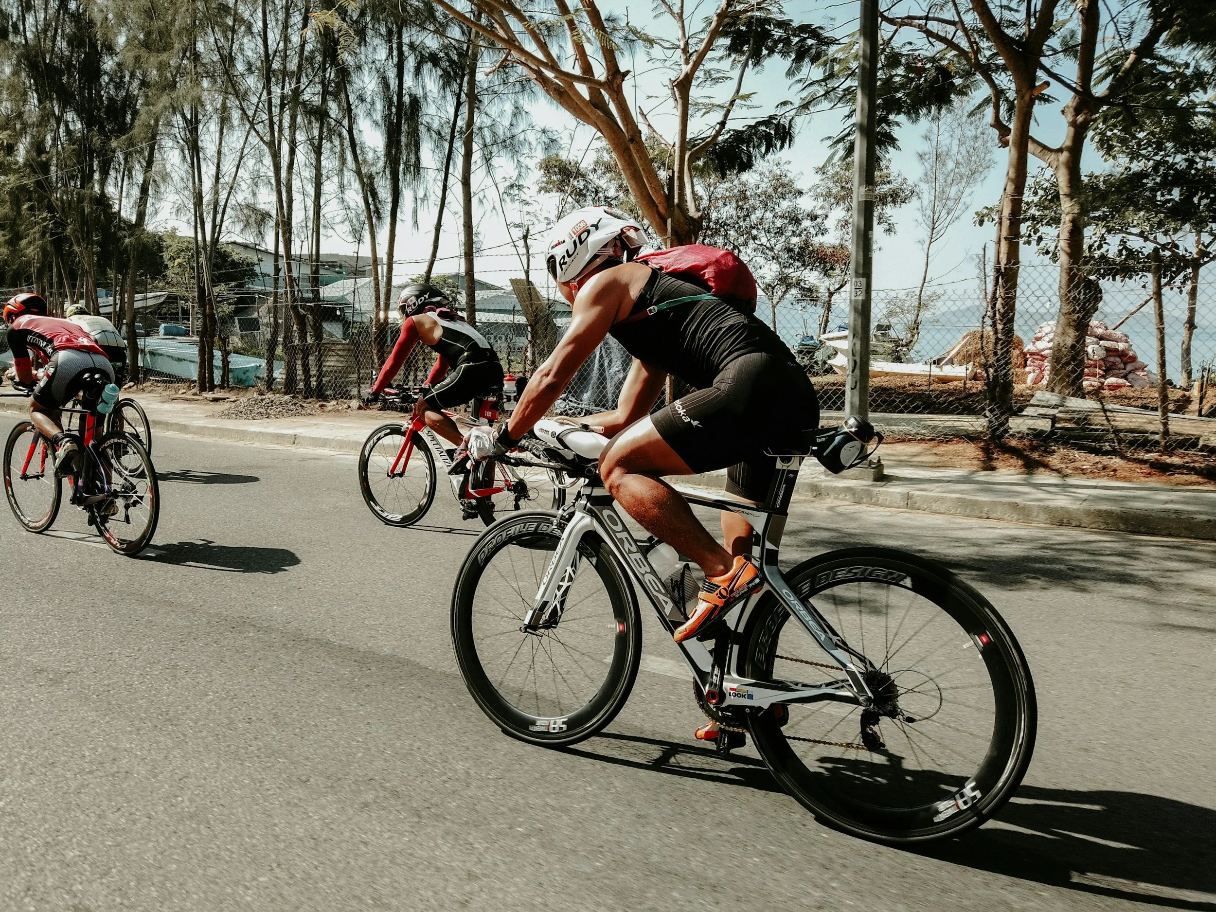 Group of cyclists riding on a paved road with trees and a fence on the side