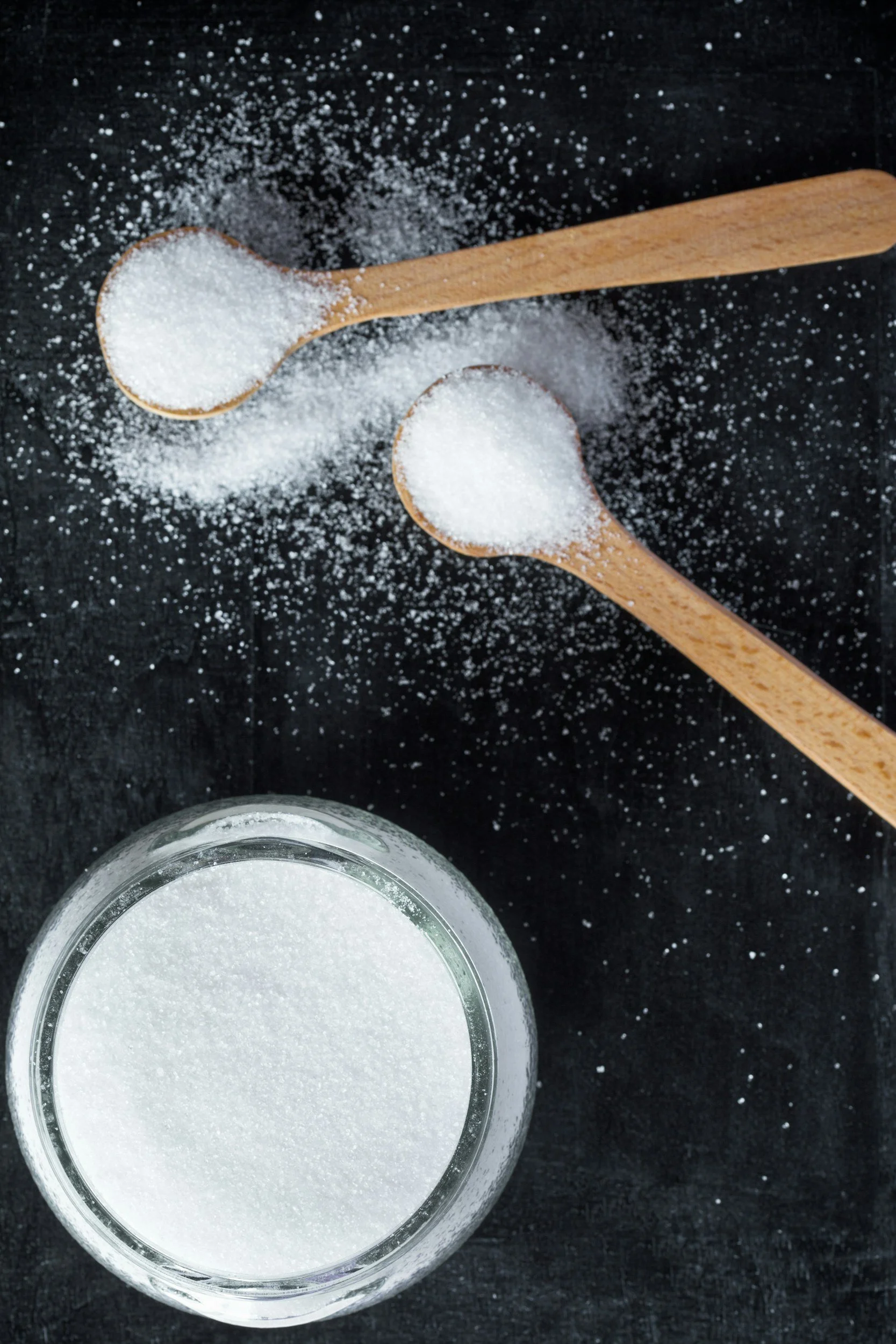 Two wooden spoons filled with white granulated sugar, with sugar spilled around them, and a glass jar filled with white sugar on a black surface.