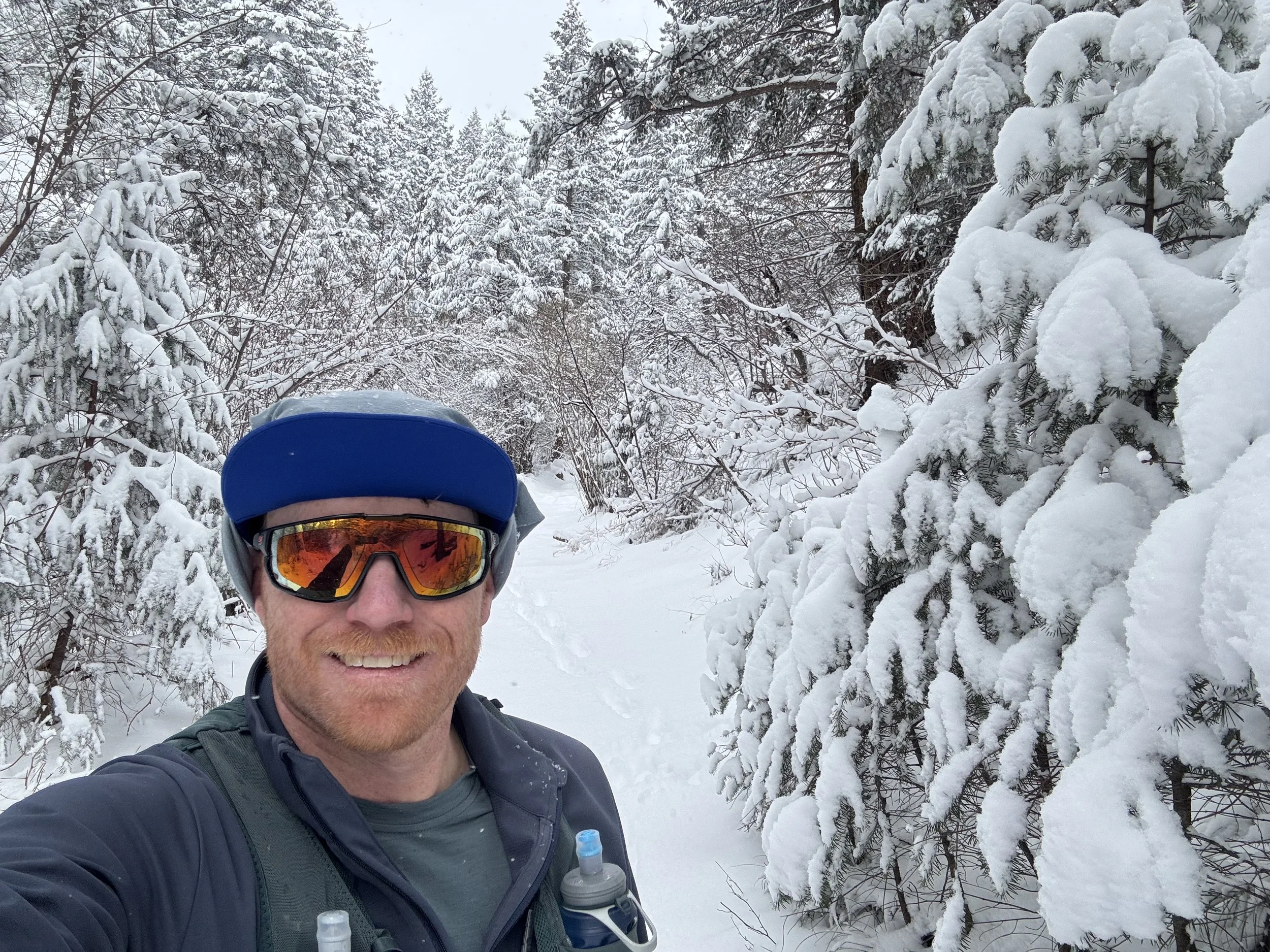 Man smiling in snow-covered forest, wearing a blue helmet, reflective sunglasses, and outdoor gear.