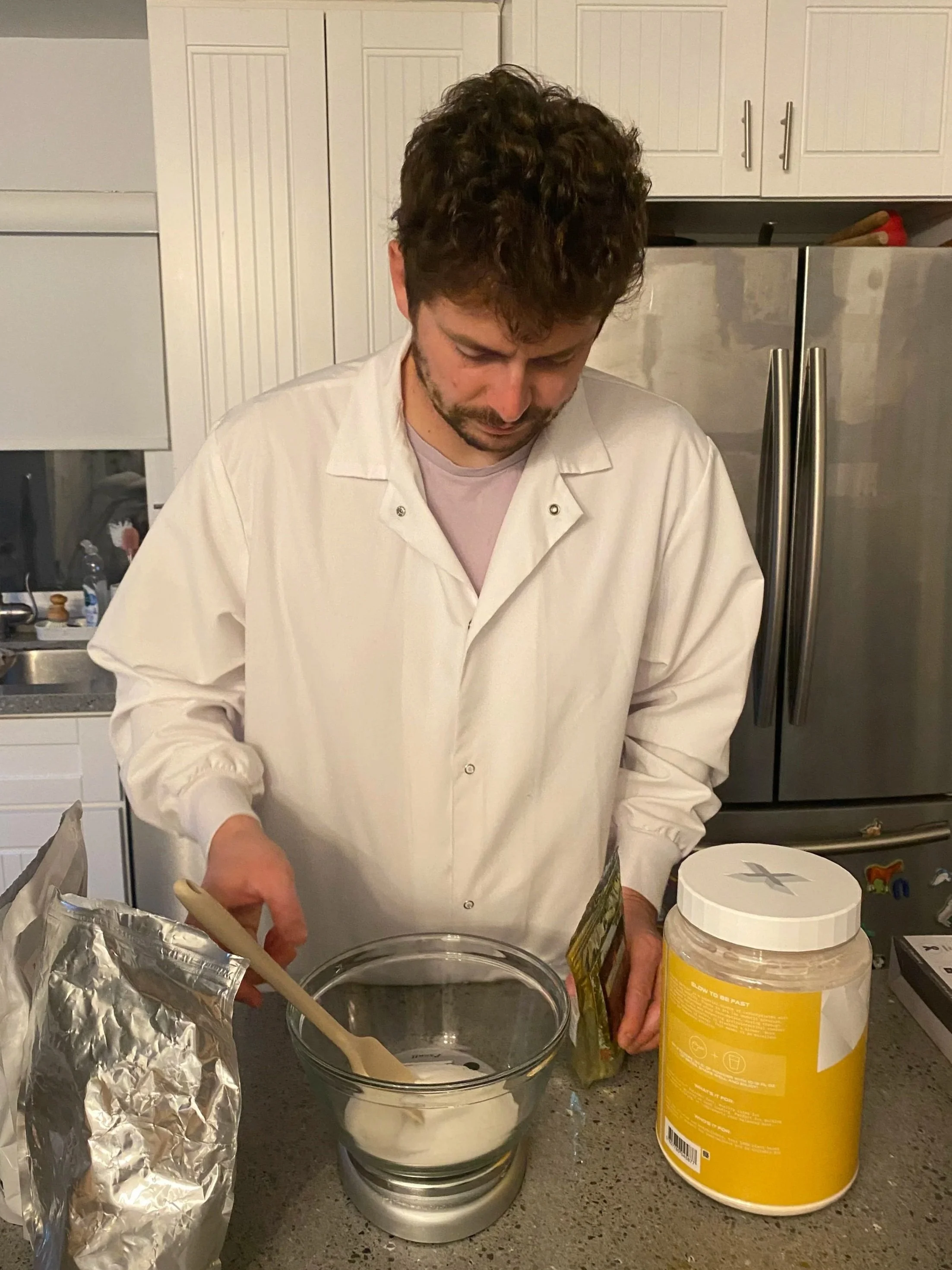 A man with curly brown hair and a beard, wearing a white lab coat, stands in a kitchen, looking down and preparing ingredients in a glass bowl on the countertop. There is a large yellow container, a package of food, and an open silver bag nearby.