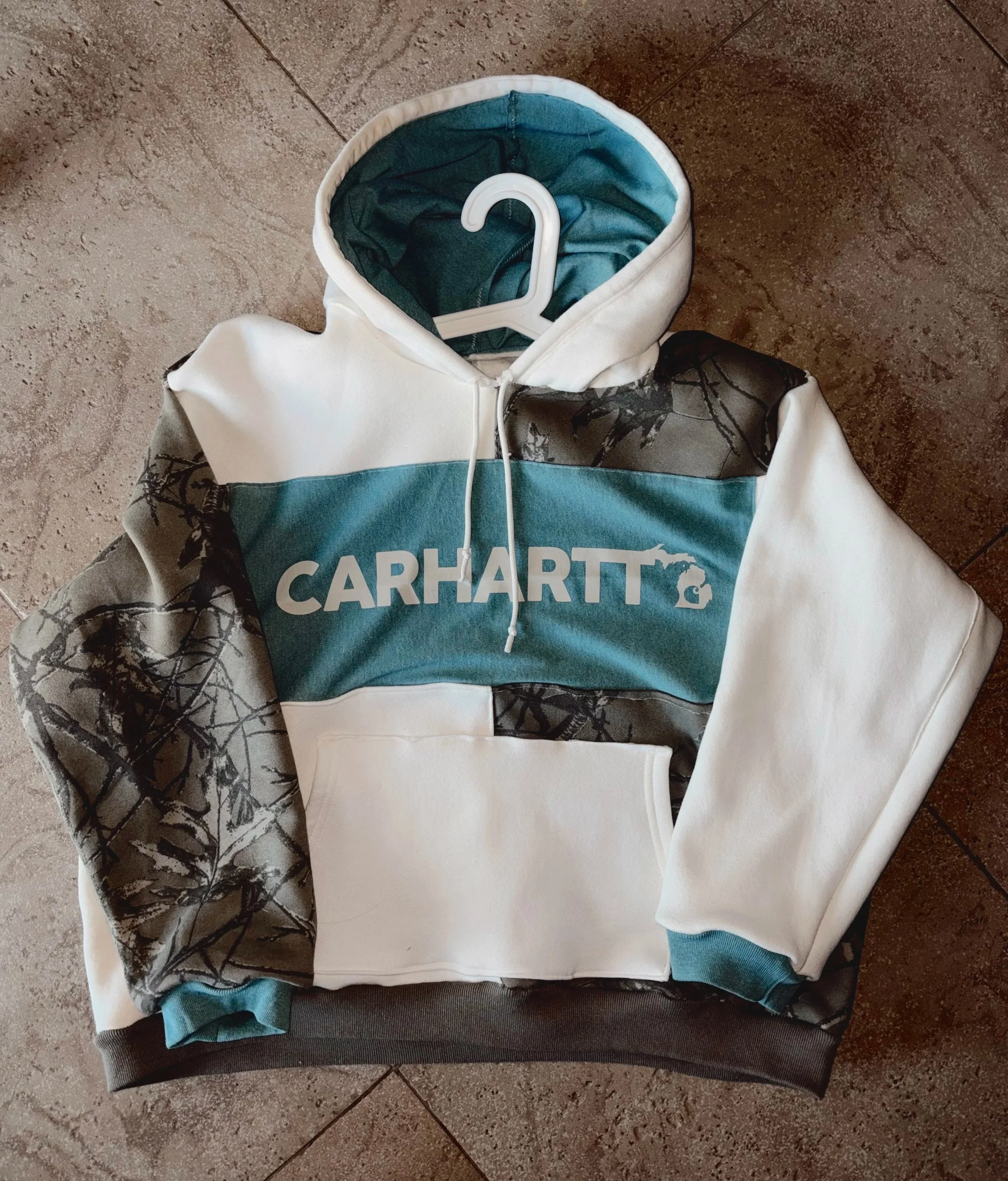 A white hooded sweatshirt with black and gray camouflage patterns on the sleeves and black accents, featuring the word 'CARHARTT' and a logo in the center of the chest.