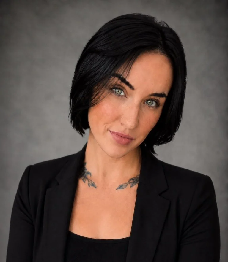Portrait of a woman with short black hair, blue eyes, wearing a black blazer and top, with tattoos on her chest, against a gray background.