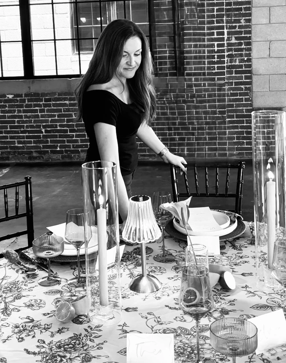 A woman standing at a table decorated for a meal, with candles and elegant tableware, in an industrial-style room with exposed brick walls.