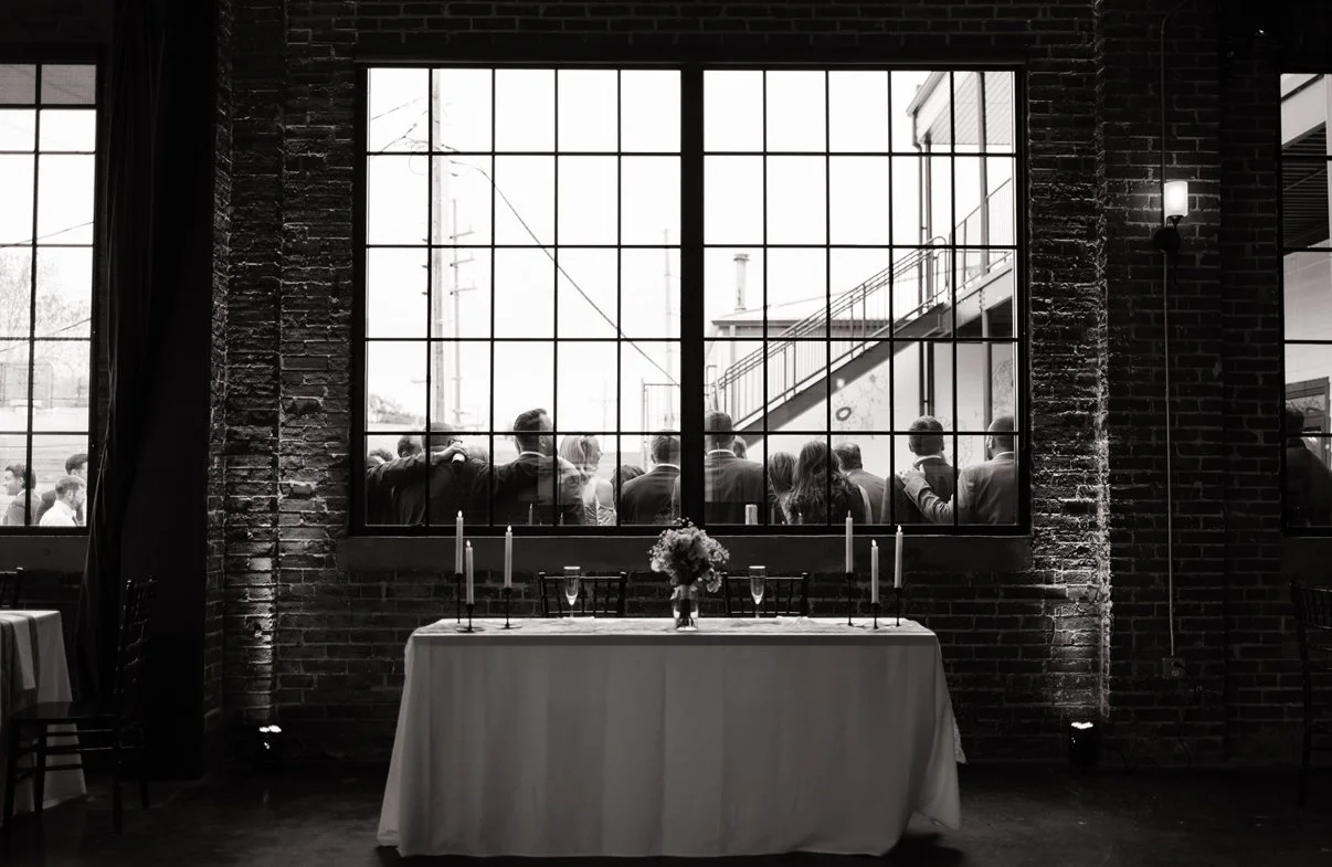Black and white photo of an indoor event space with a long table set with candles and flowers in front of large industrial-style windows. Outside the window, a group of people can be seen socializing.