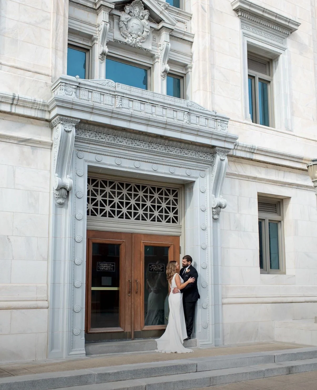 A bride and groom standing close together outside a historic building with detailed stone architecture, double wooden doors, and large windows.