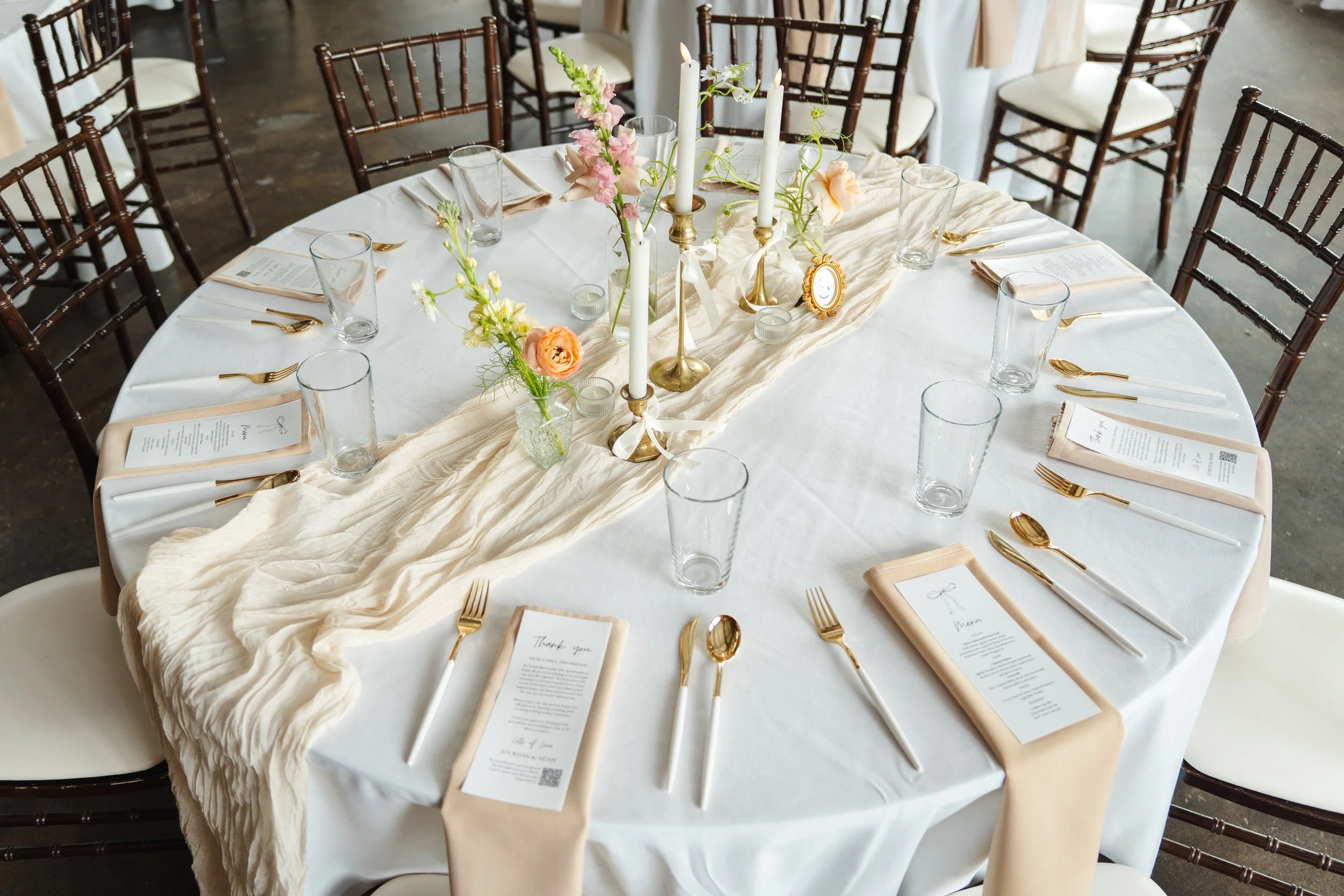 Round wedding reception table with white tablecloth, beige runner, floral centerpiece, candles, gold cutlery, glasses, and menus.
