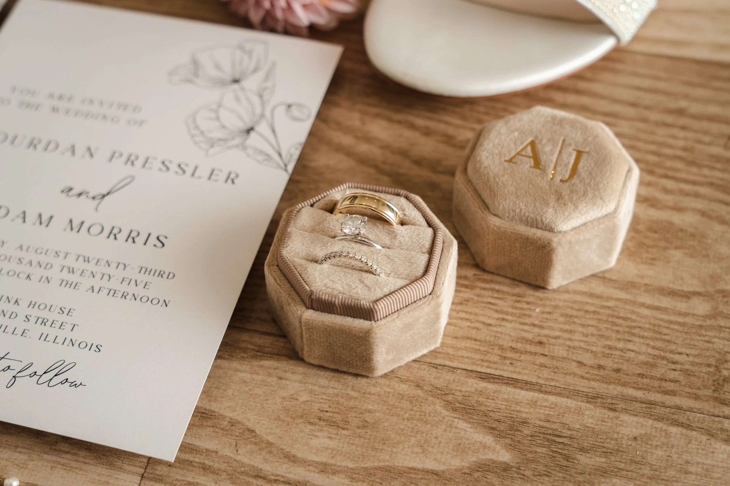 A velvet ring box with a gold wedding band, a diamond ring, and a delicate band inside, placed on a wooden surface next to a matching velvet box with the initials 'A.J.' in gold, and a wedding invitation with floral illustrations.