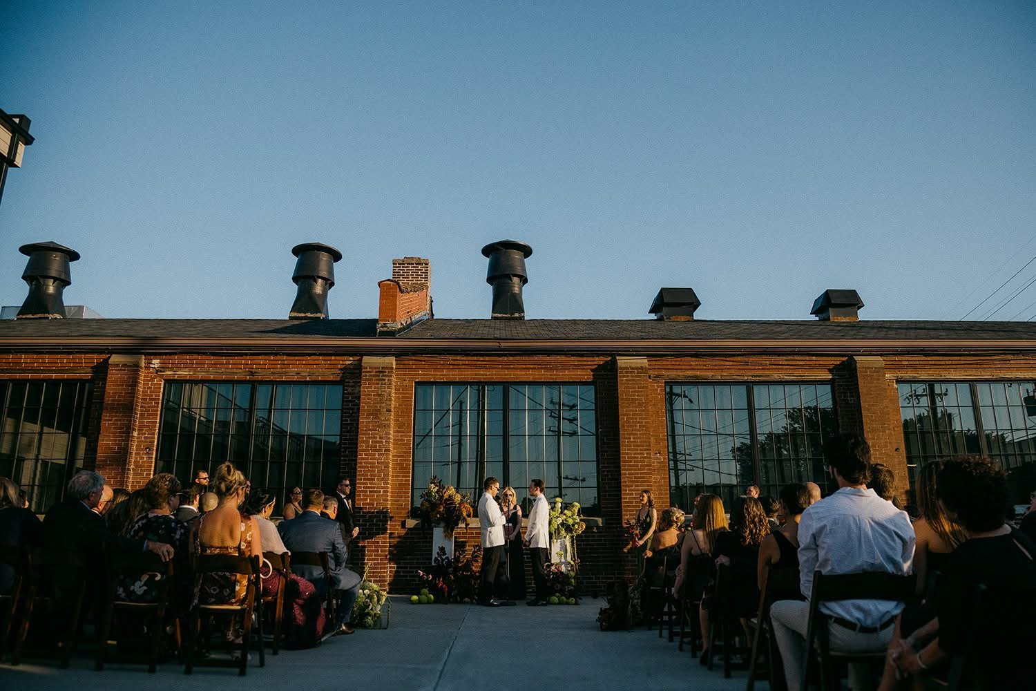 Outdoor wedding ceremony with guests seated in front of a brick building with large windows, and a couple standing before officiants under a clear sky.