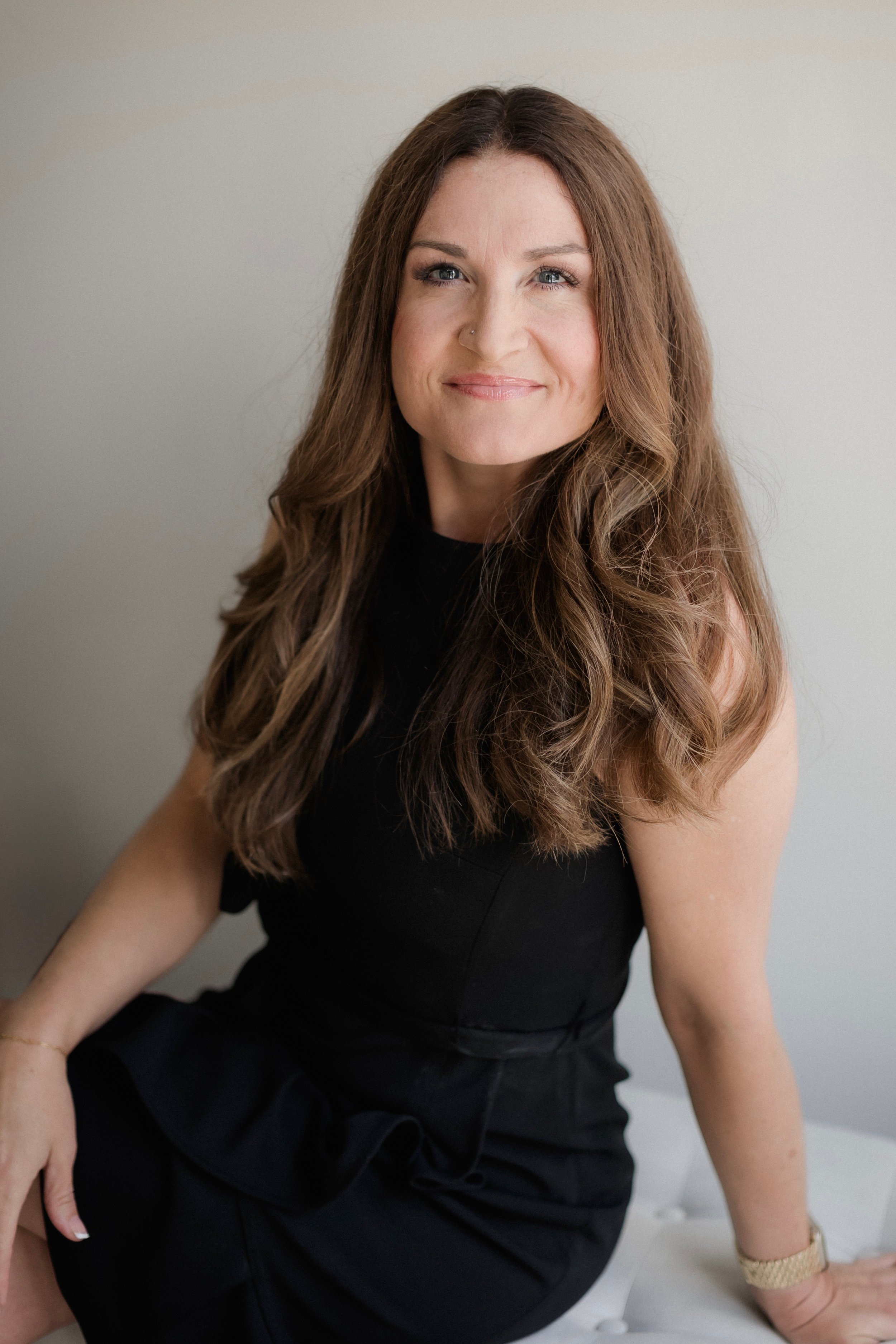 A woman with long wavy brown hair, light blue eyes, and a nose piercing, wearing a black sleeveless dress, sitting on a white surface against a plain light-colored wall, smiling softly.