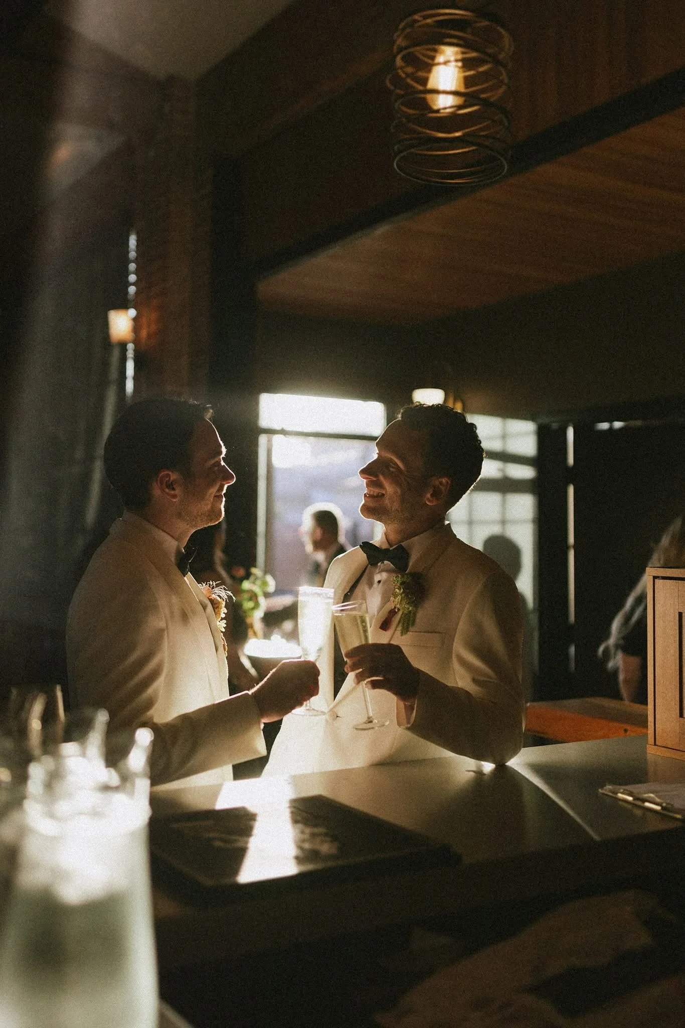 Two men in tuxedos sharing a toast and smiling at each other in a dimly lit restaurant or bar, with sunlight streaming through the windows behind them.