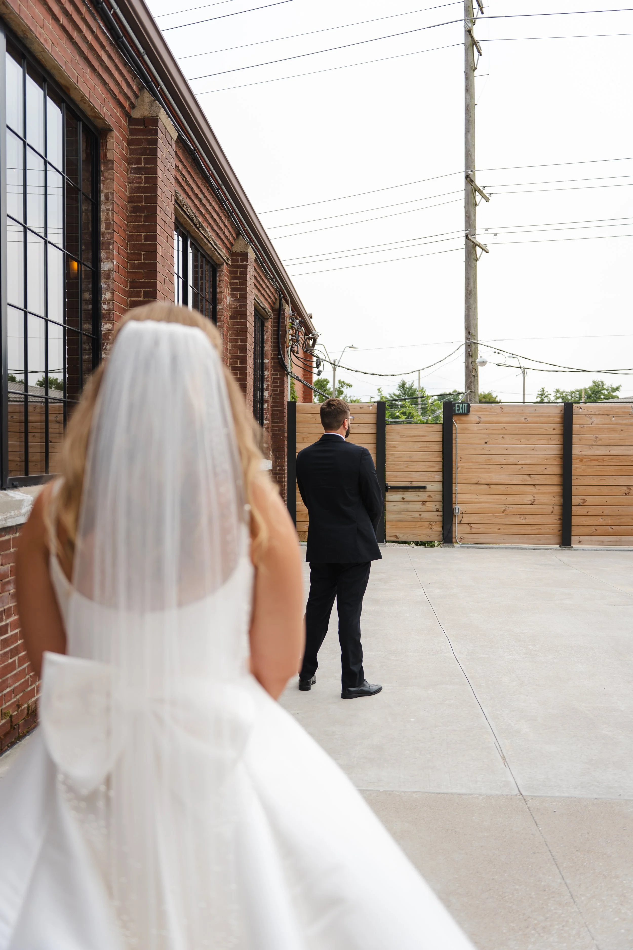 A bride wearing a wedding dress and veil watches a groom in a suit standing apart outside near a brick building and wooden fence.