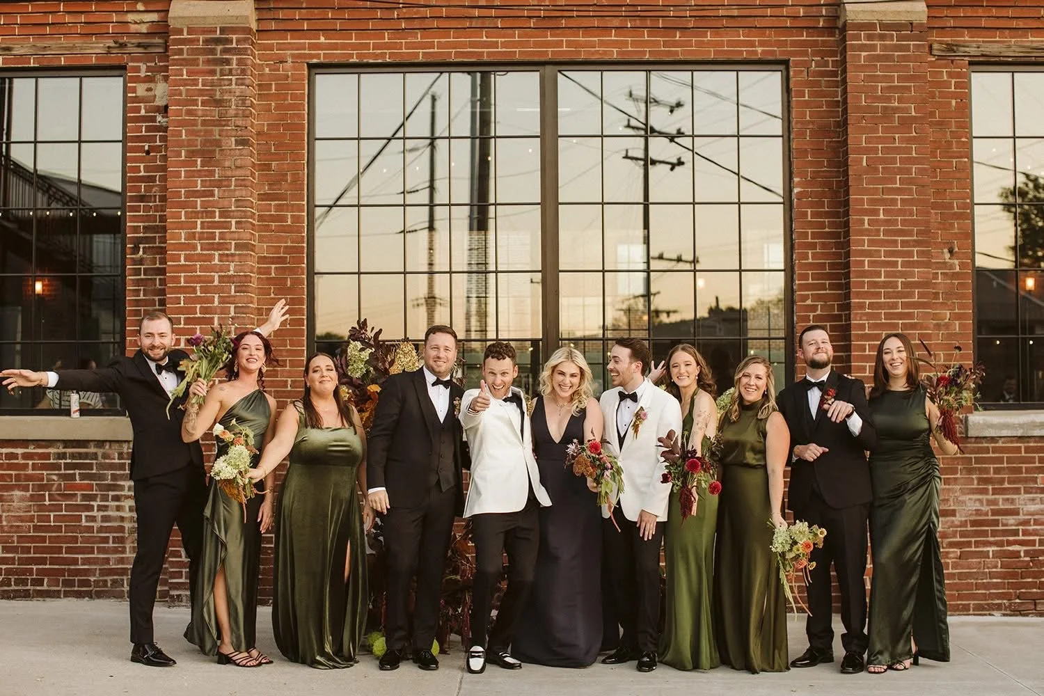 Group of wedding party members dressed in formal attire standing outside in front of a large brick building with big glass windows, holding bouquets and celebrating.