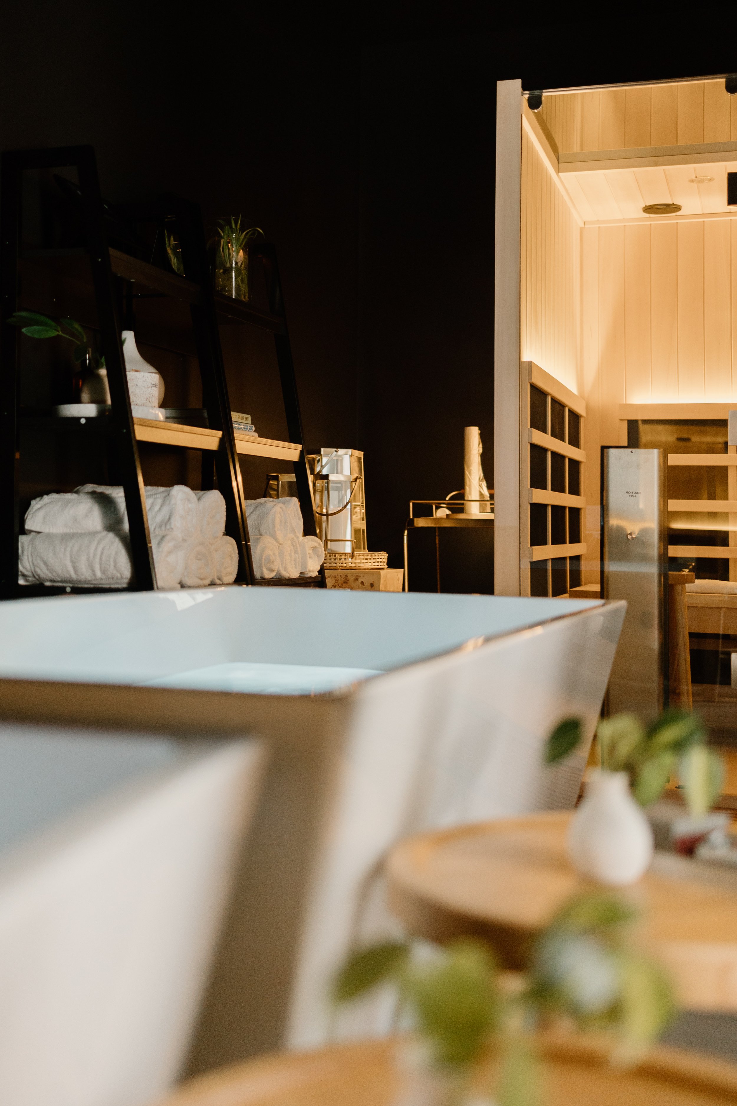 Interior of a spa or wellness center with a white bathtub in the foreground, wooden shelves with rolled towels, and a sauna room in the background.