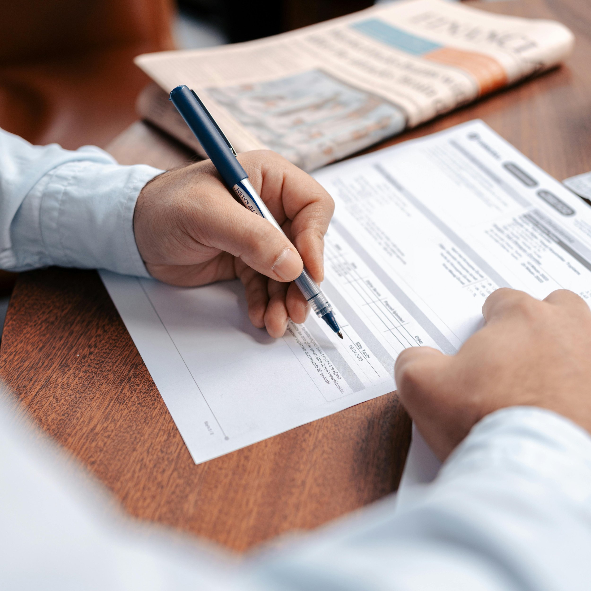 Person filling out a form with a blue pen, with a newspaper on the table.