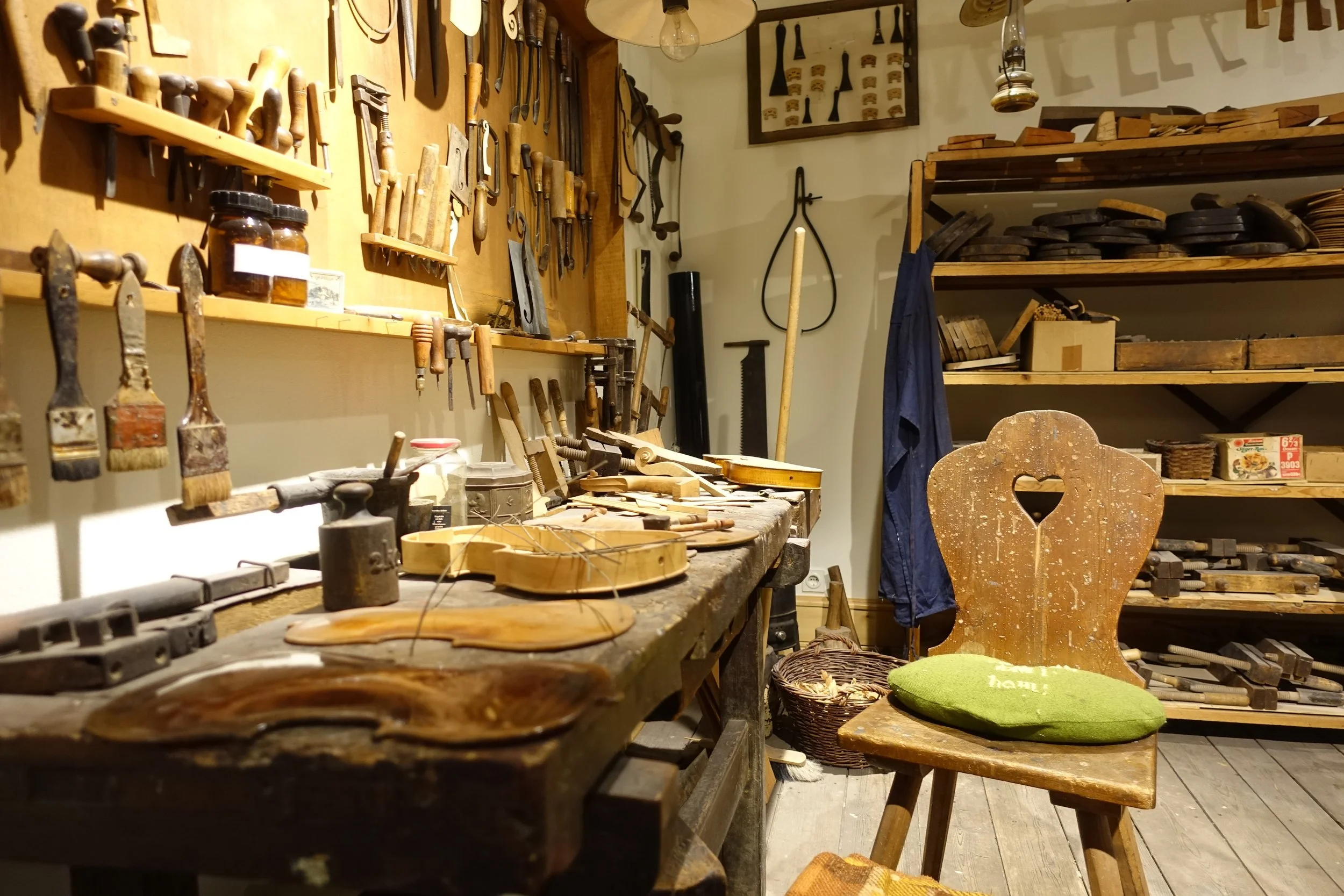 A woodworking workshop with a cluttered workbench, assorted hand tools on the wall, and shelves filled with wooden objects and materials. A wooden chair with a green cushion is in the foreground.