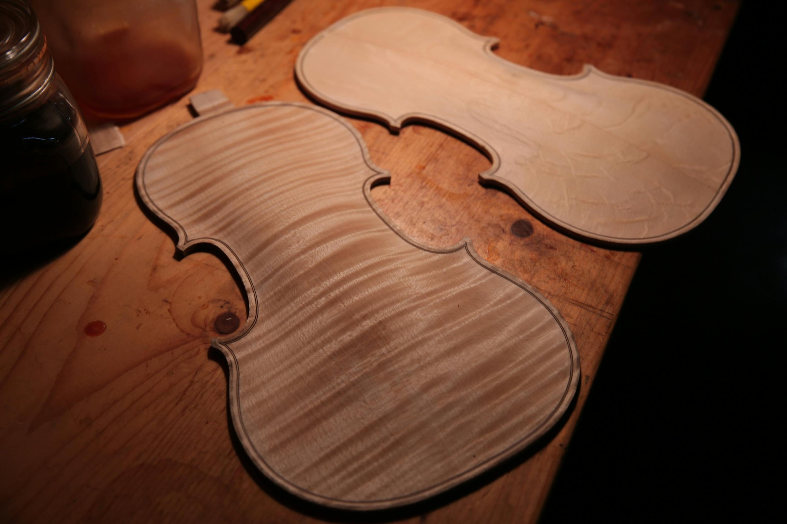 Two unassembled wooden violin bodies on a workbench, surrounded by woodworking tools and supplies.