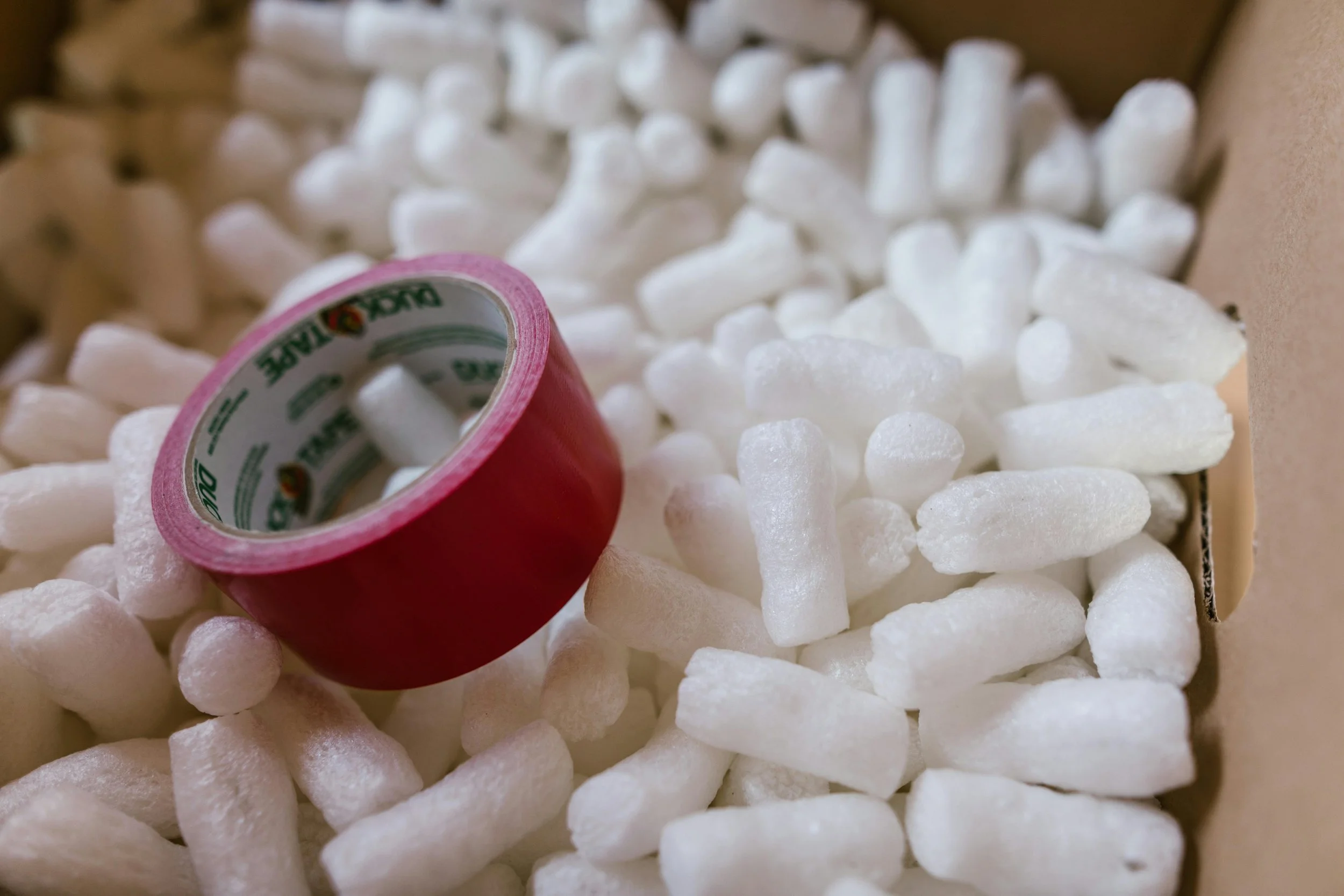 A close-up of packing peanuts and a roll of pink tape in a cardboard box.