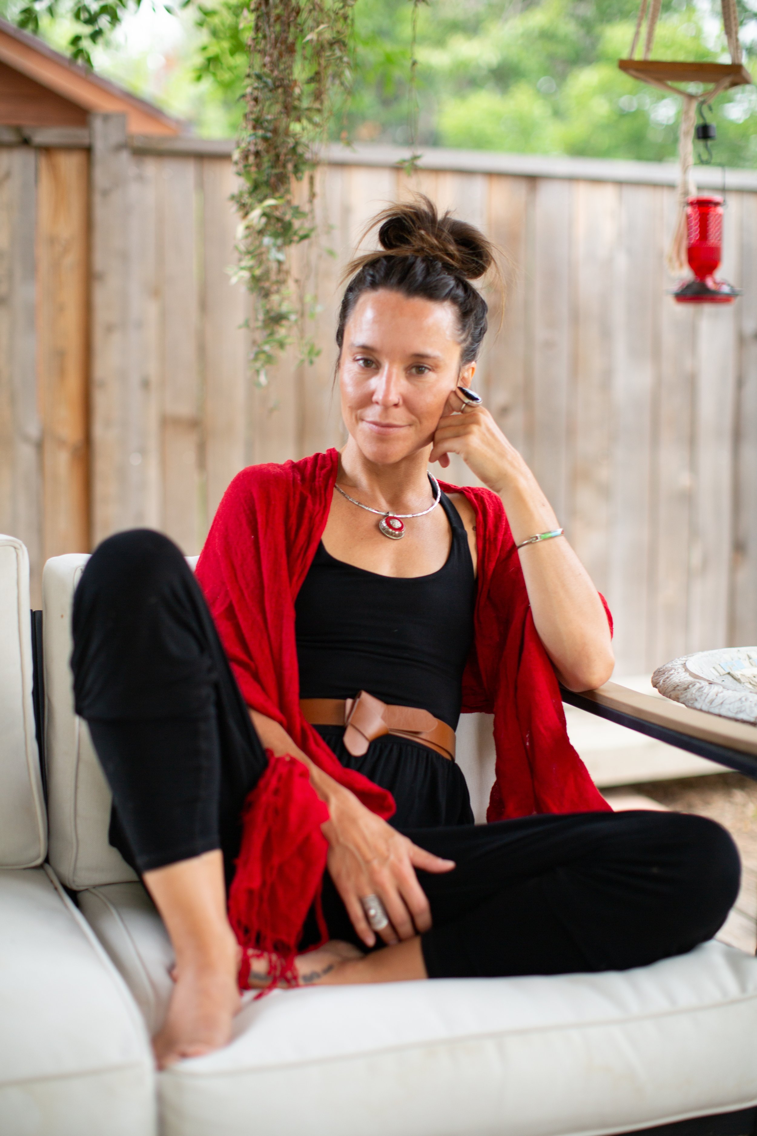 A woman with a messy bun, wearing a red cardigan and jewelry, is sitting on a white cushioned outdoor seating area. She has a relaxed expression and is touching her face with her right hand, with a wooden fence and trees in the background.