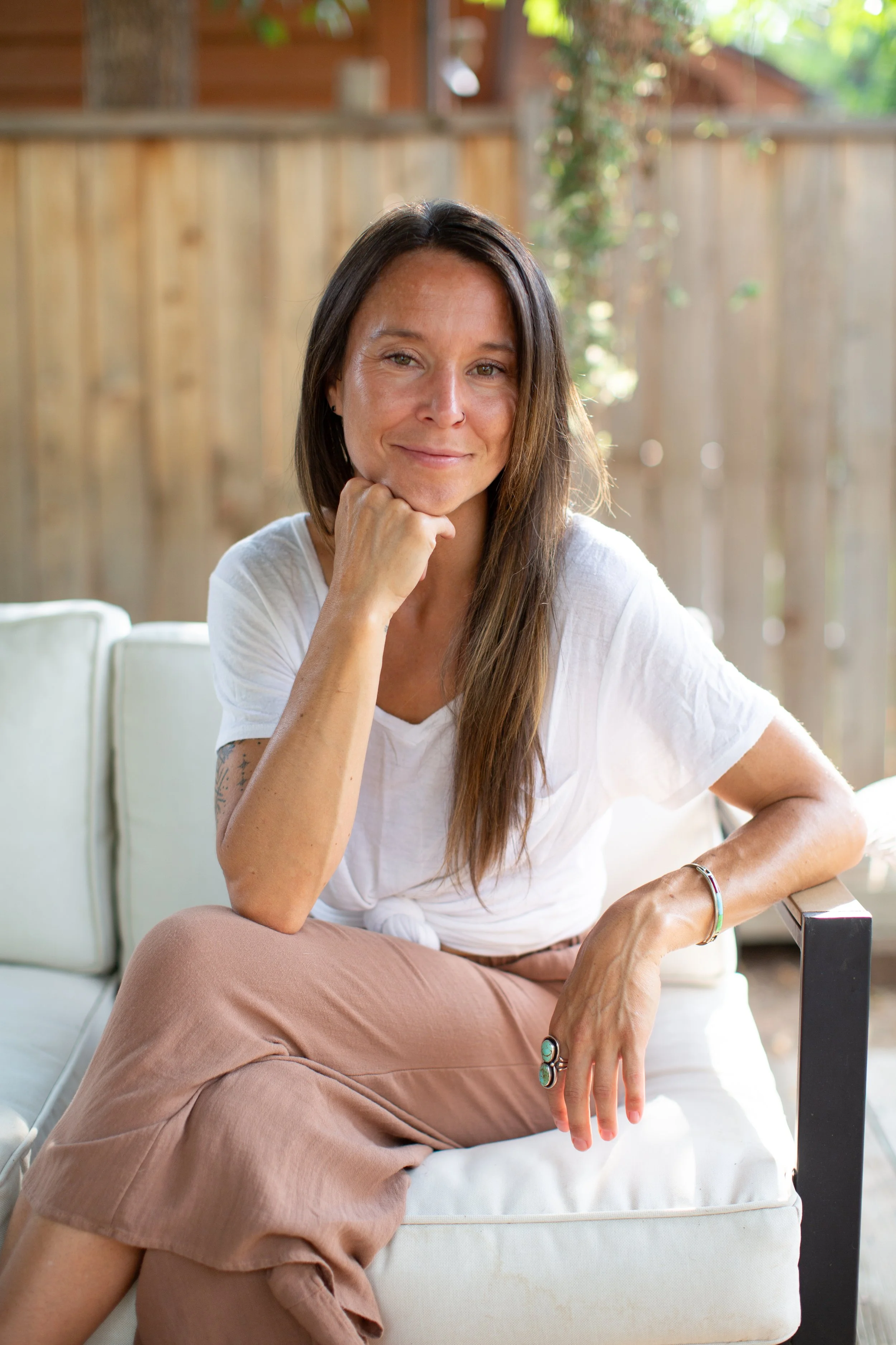 A woman with long brown hair sitting outdoors near a wooden fence, wearing a white t-shirt and beige pants, smiling slightly, resting her chin on her hand.