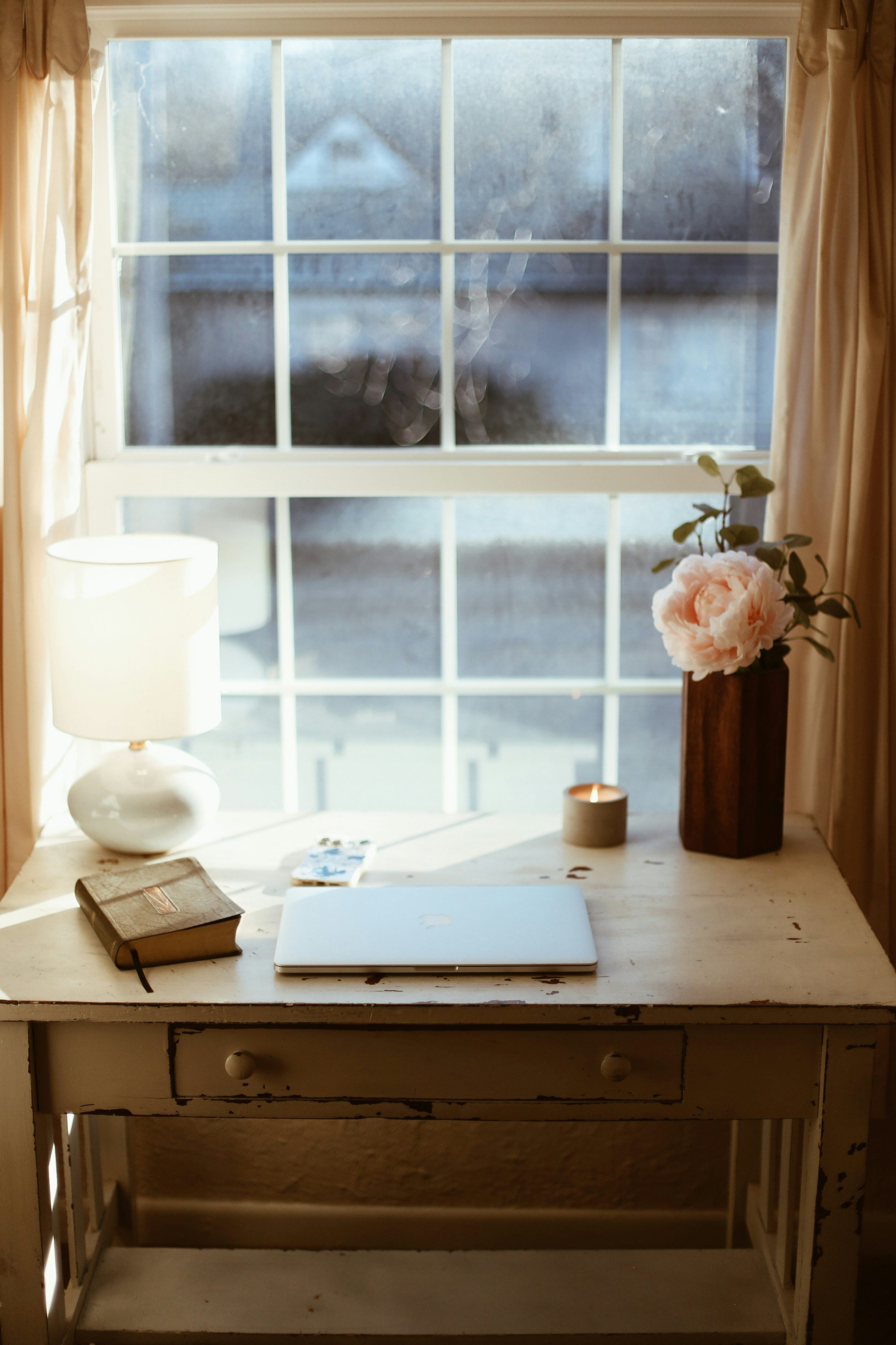A white distressed wooden desk near a window with beige curtains, containing a white table lamp, an old book, a closed laptop, a candle, a vase with pink flowers, and a few small items.