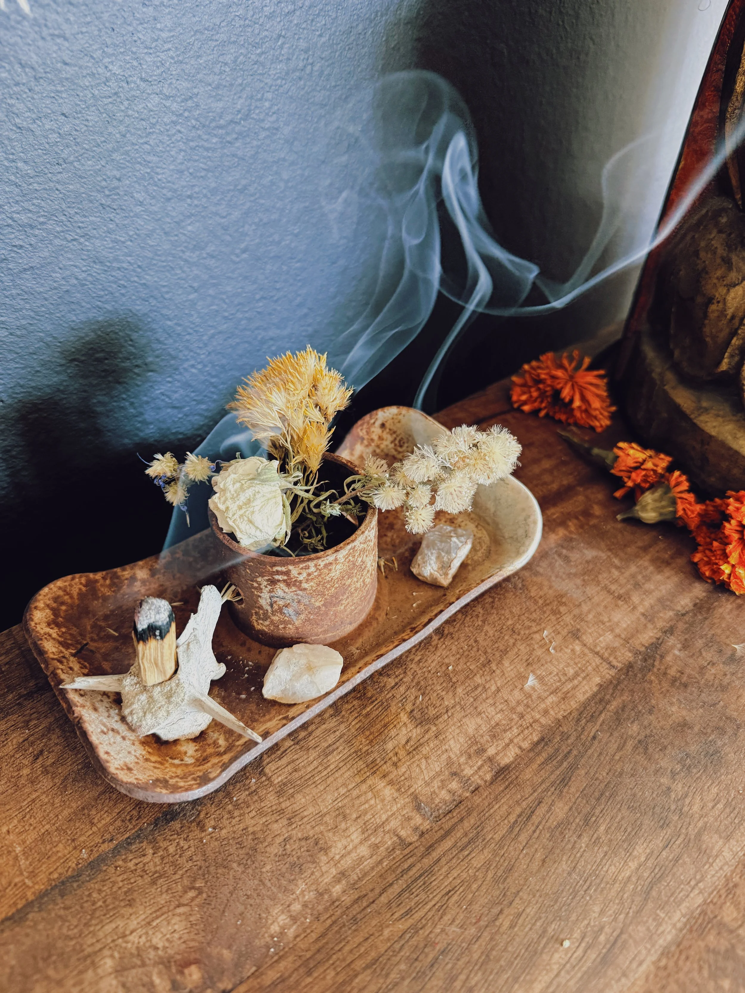A wooden tray on a table holds dried flowers, stones, and a piece of coral, with a wall and some dried flowers in the background.