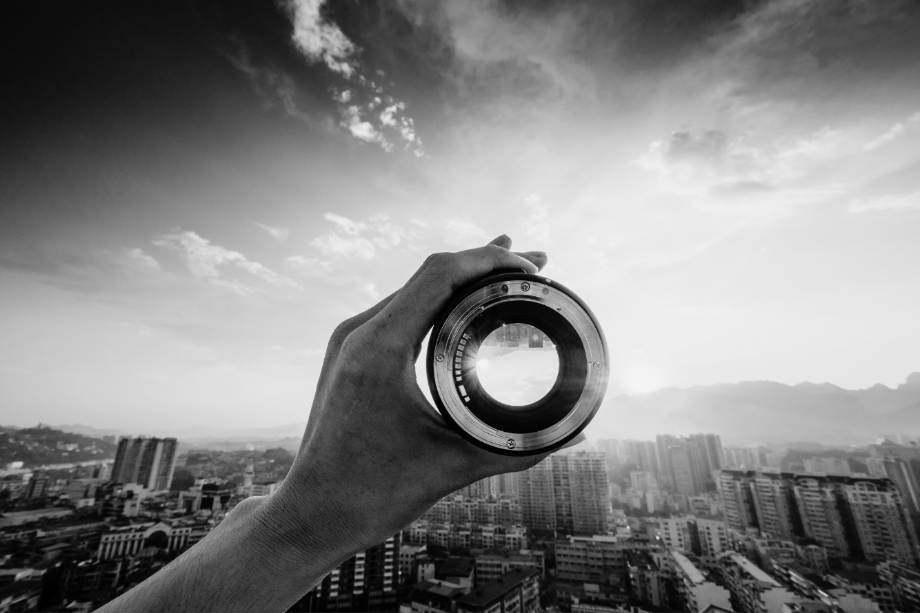 A black and white photo of a hand holding a camera lens with a cityscape and mountains in the background.