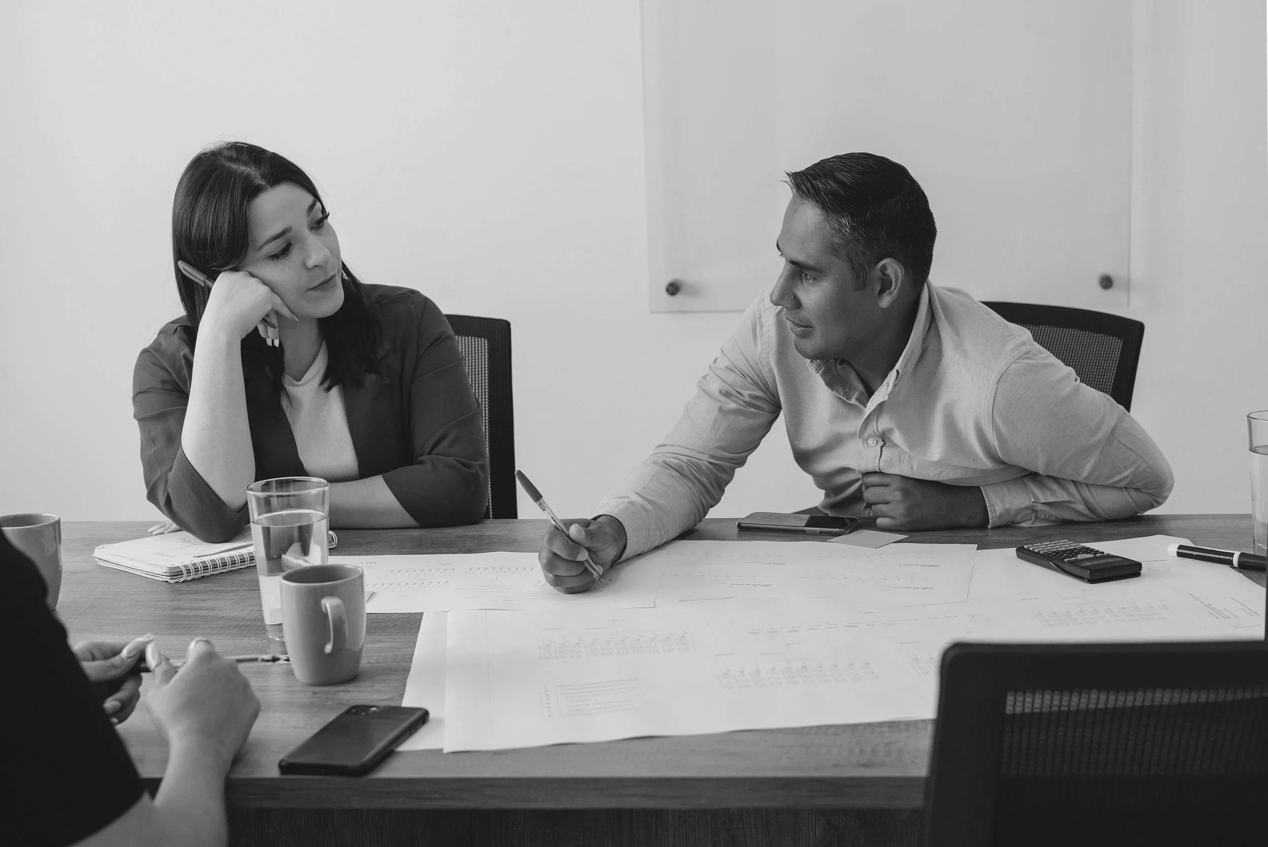 Two people in a meeting room with documents, notebooks, cups, and objects on the table, engaged in a conversation. One woman appears to be listening thoughtfully, while a man speaks and gestures with a pen.
