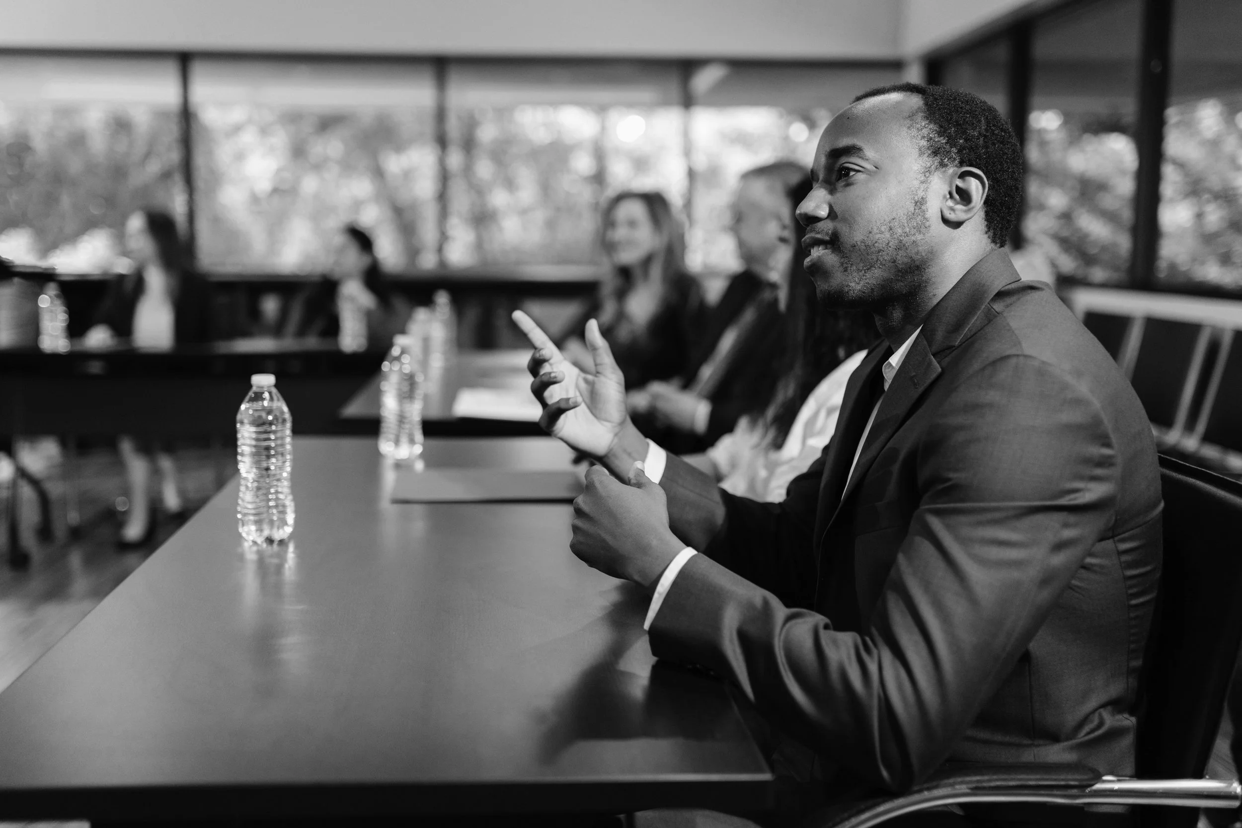 A man in a suit and tie is sitting at a conference table, speaking or explaining something during a meeting. There are water bottles and other people sitting at the table with him. The background shows windows with trees outside.