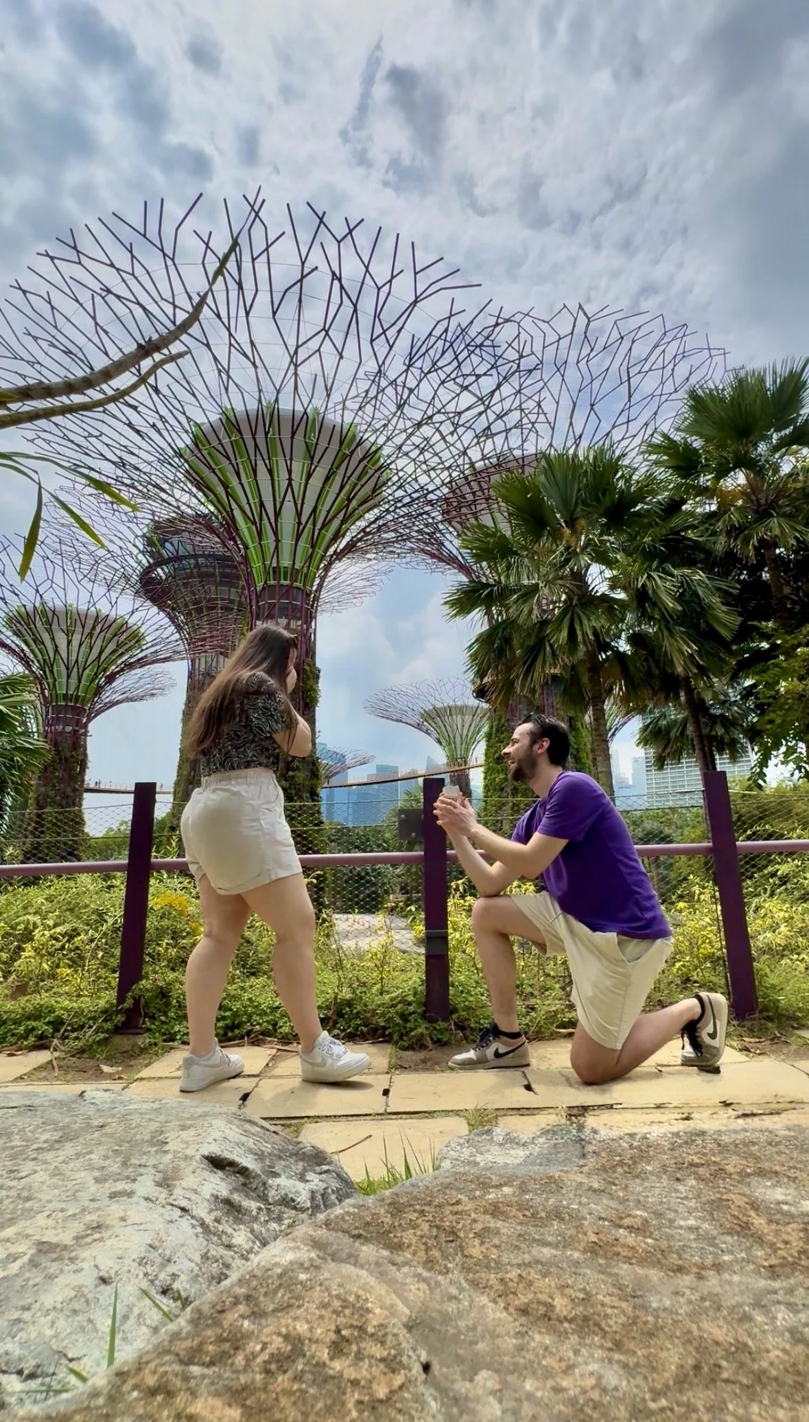 Een man doet een voorstel voor een vriendin in een park met grote, metalen boomstructuren en groen.