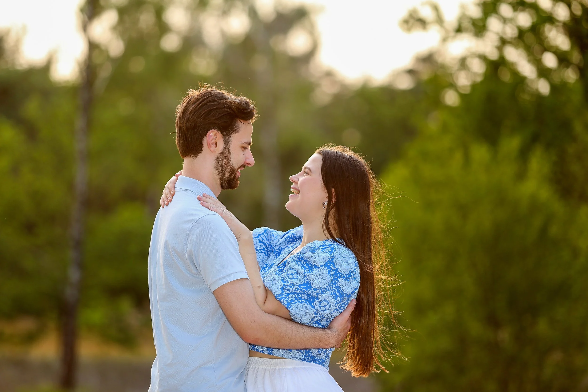 Een man en vrouw die lachen en elkaar vasthouden in een bos met groene bomen op de achtergrond.