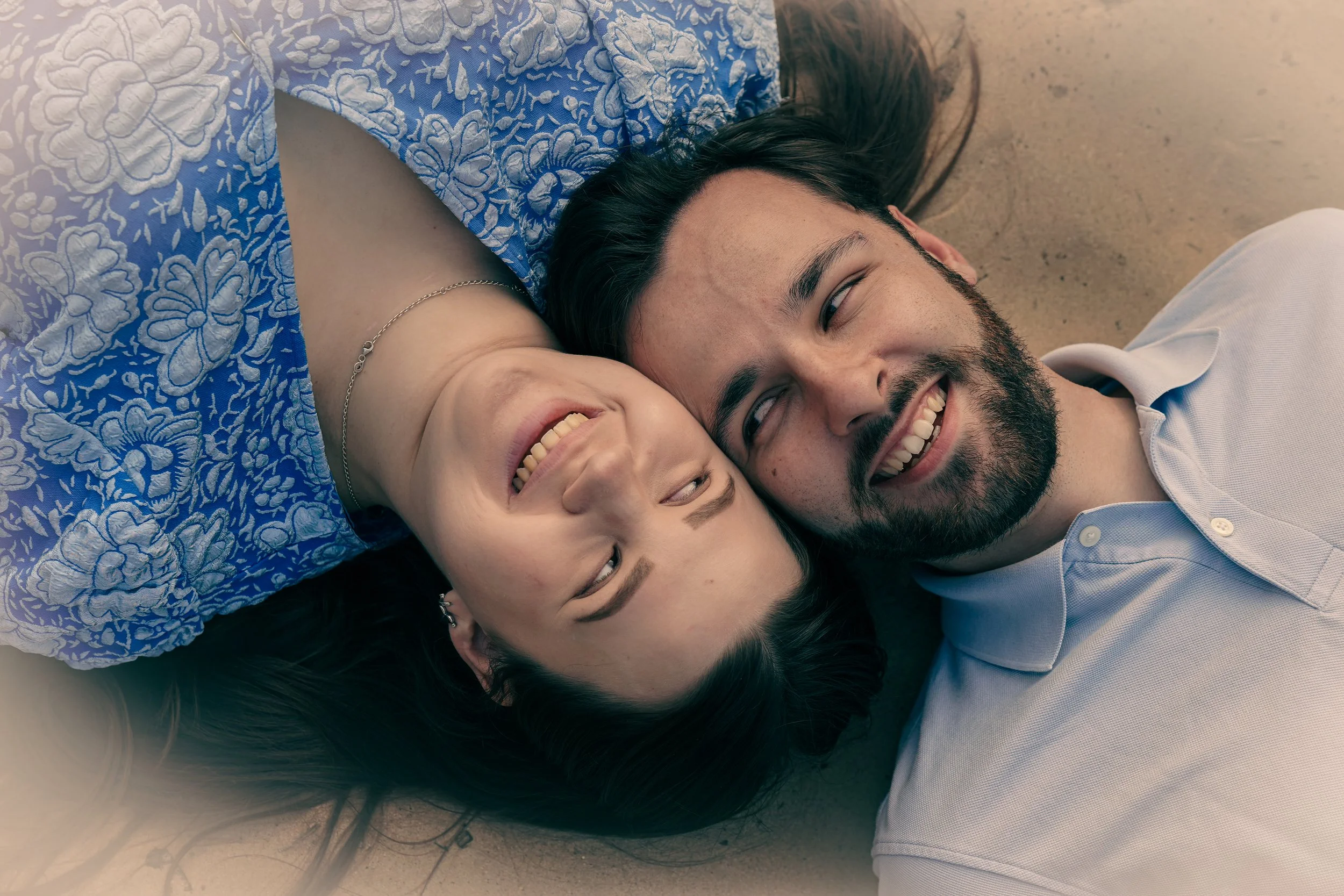 Twee mensen lachen terwijl ze samen op de grond liggen, een vrouw met een blauwe jurk en een man met een witte polo.
