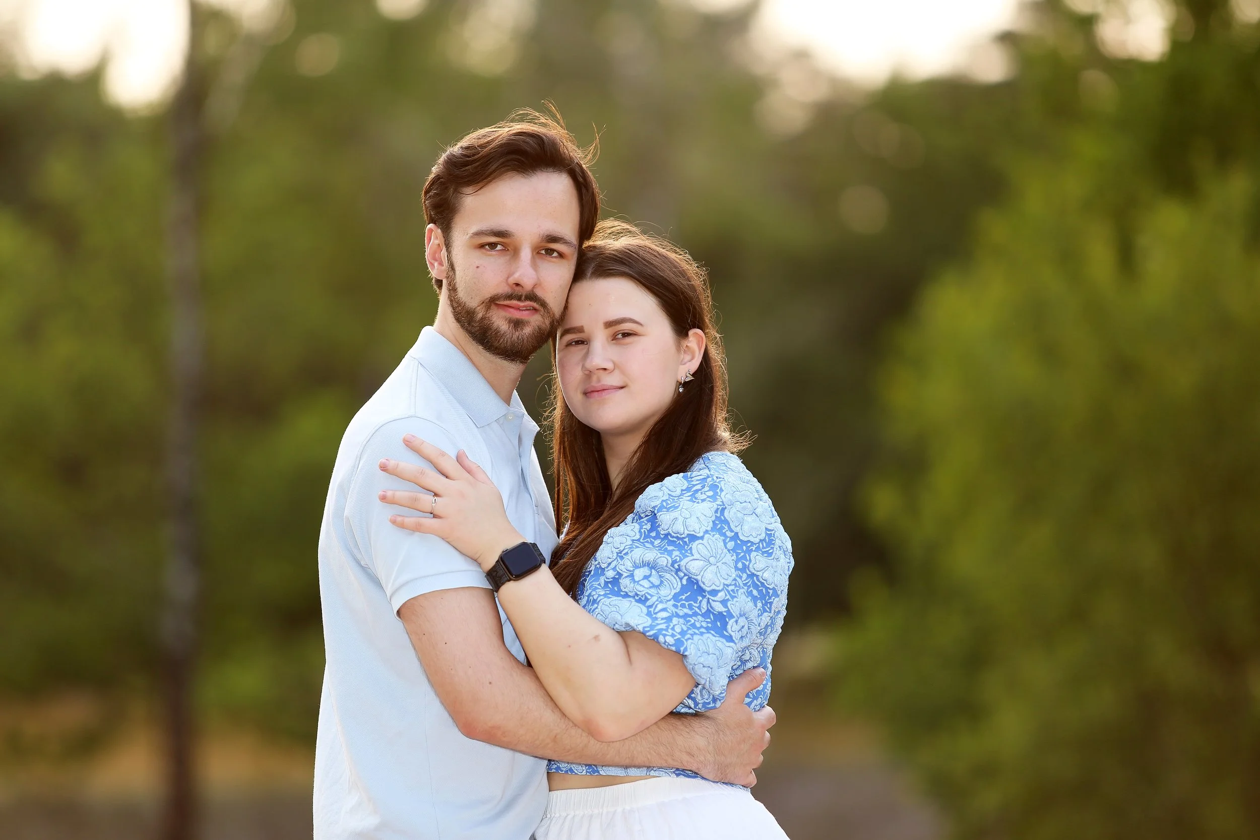 Een jonge man en vrouw omhelzen elkaar in een bos, de man draagt een wit shirt en de vrouw een blauwe jurk met bloemen.
