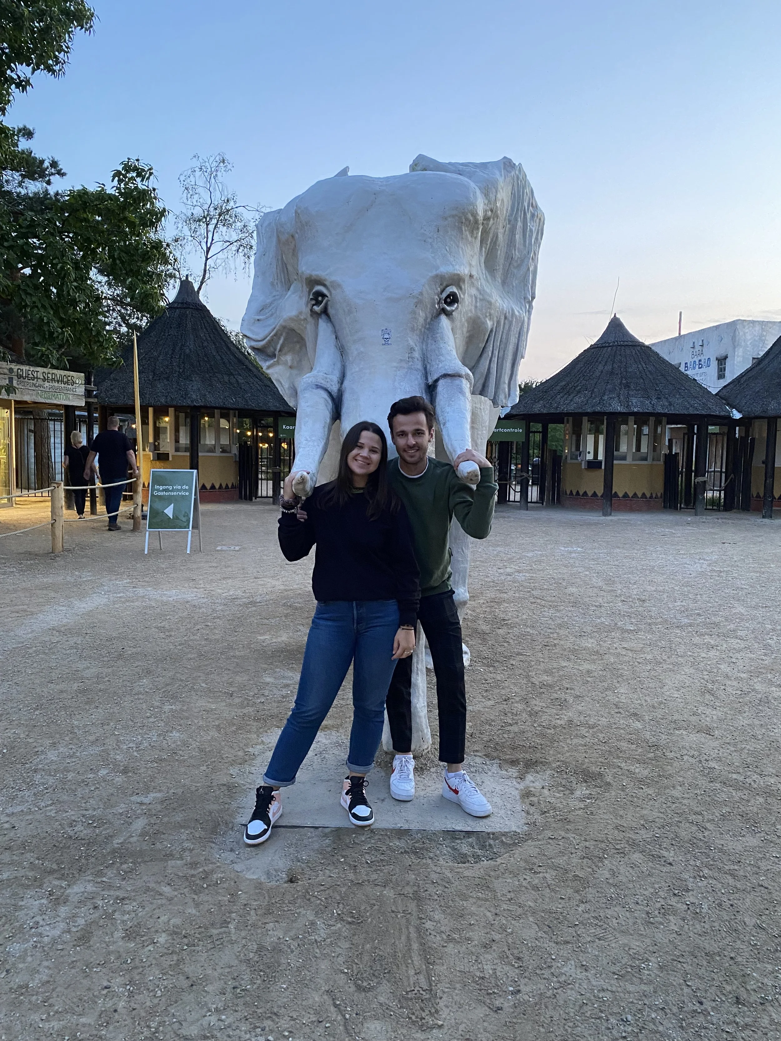 Twee mensen staan voor een nijlpaardstandbeeld in een dierentuin, 's avonds met schemerlicht, omringd door toeristen en gebouwen.