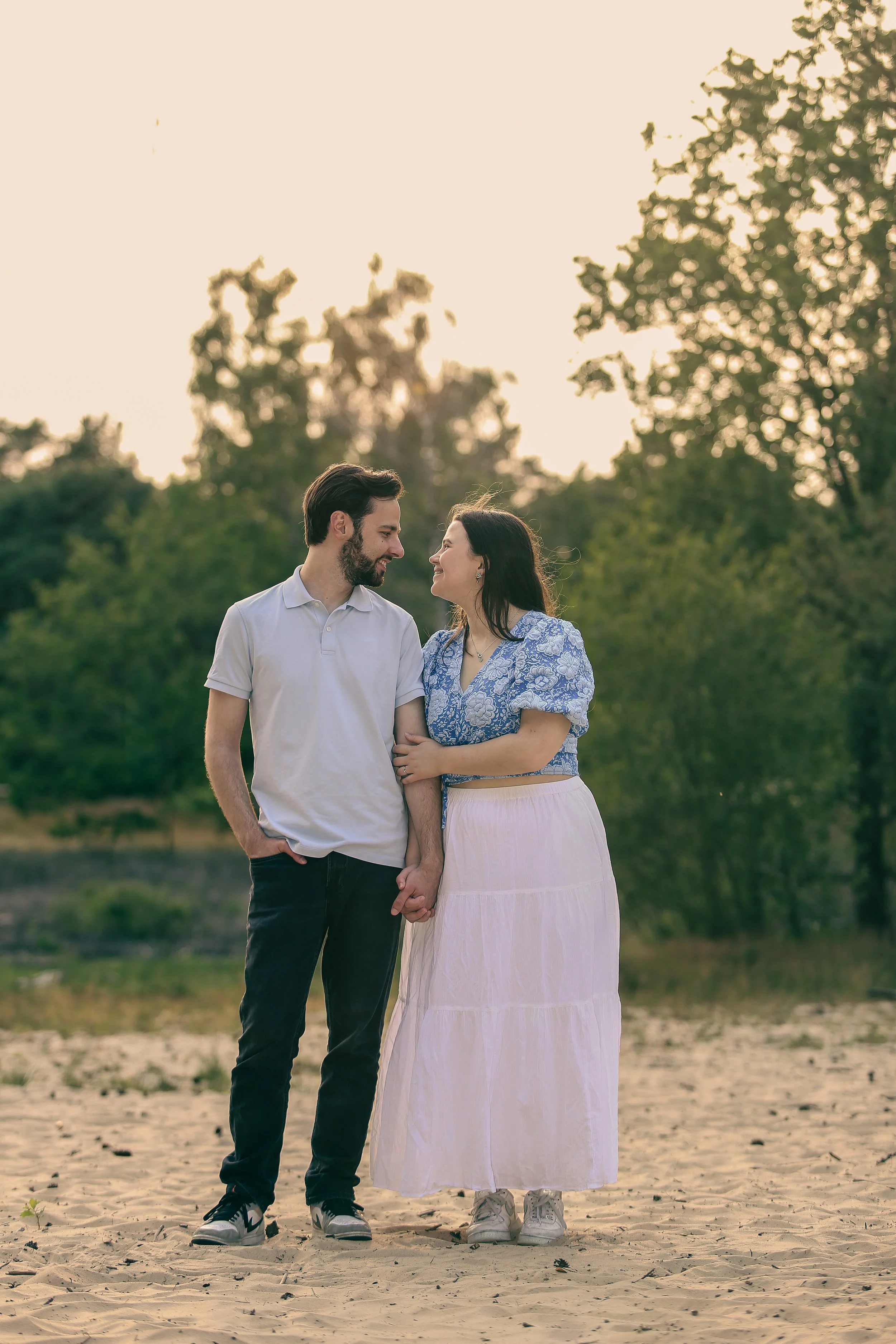 Een stel dat hand in hand staat op een zandstrand, omgeven door groene bomen, tijdens zonsondergang.