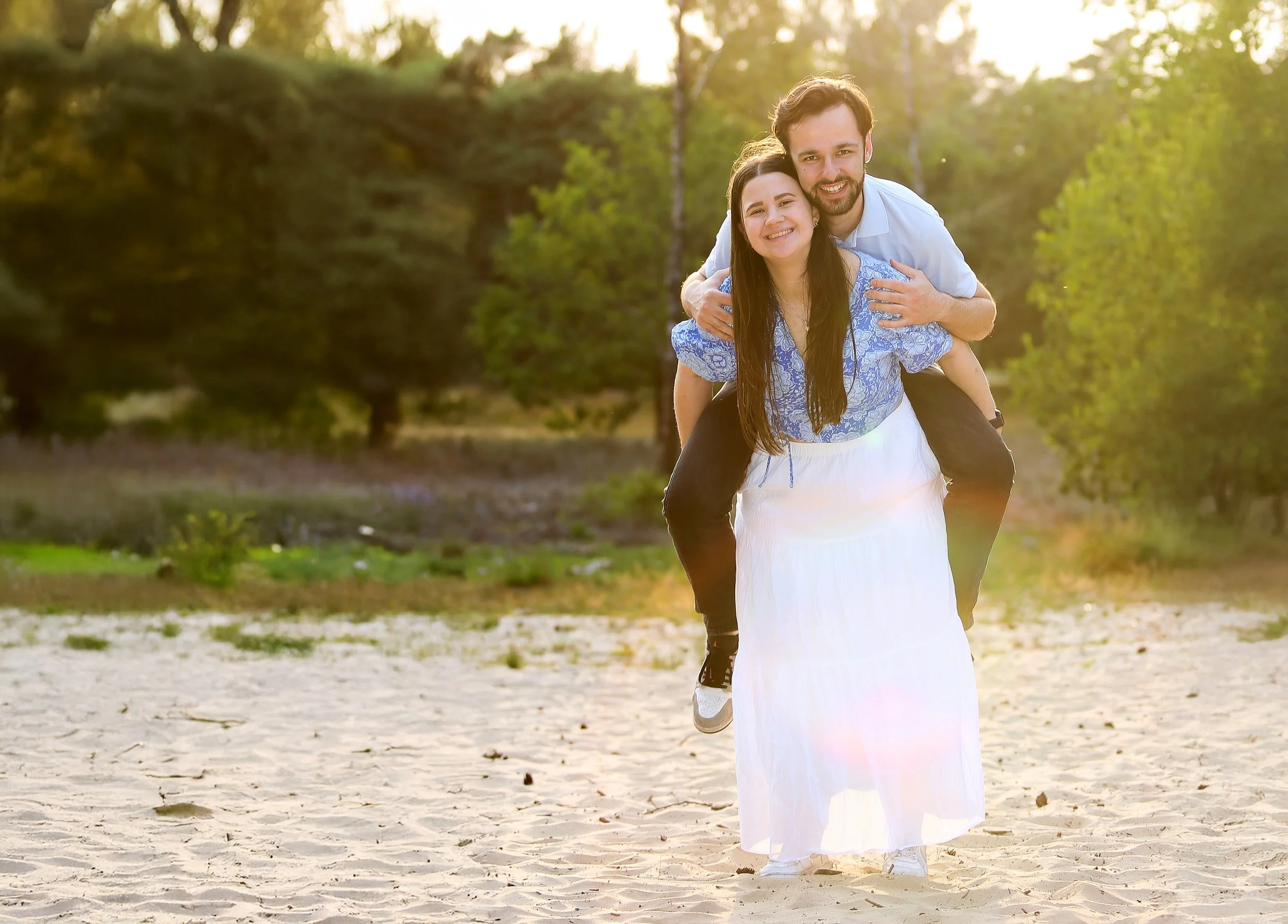 Een lachend paar op een zandstrand, de man geeft de vrouw een ritje op zijn rug, met groene bomen op de achtergrond bij zonsondergang.