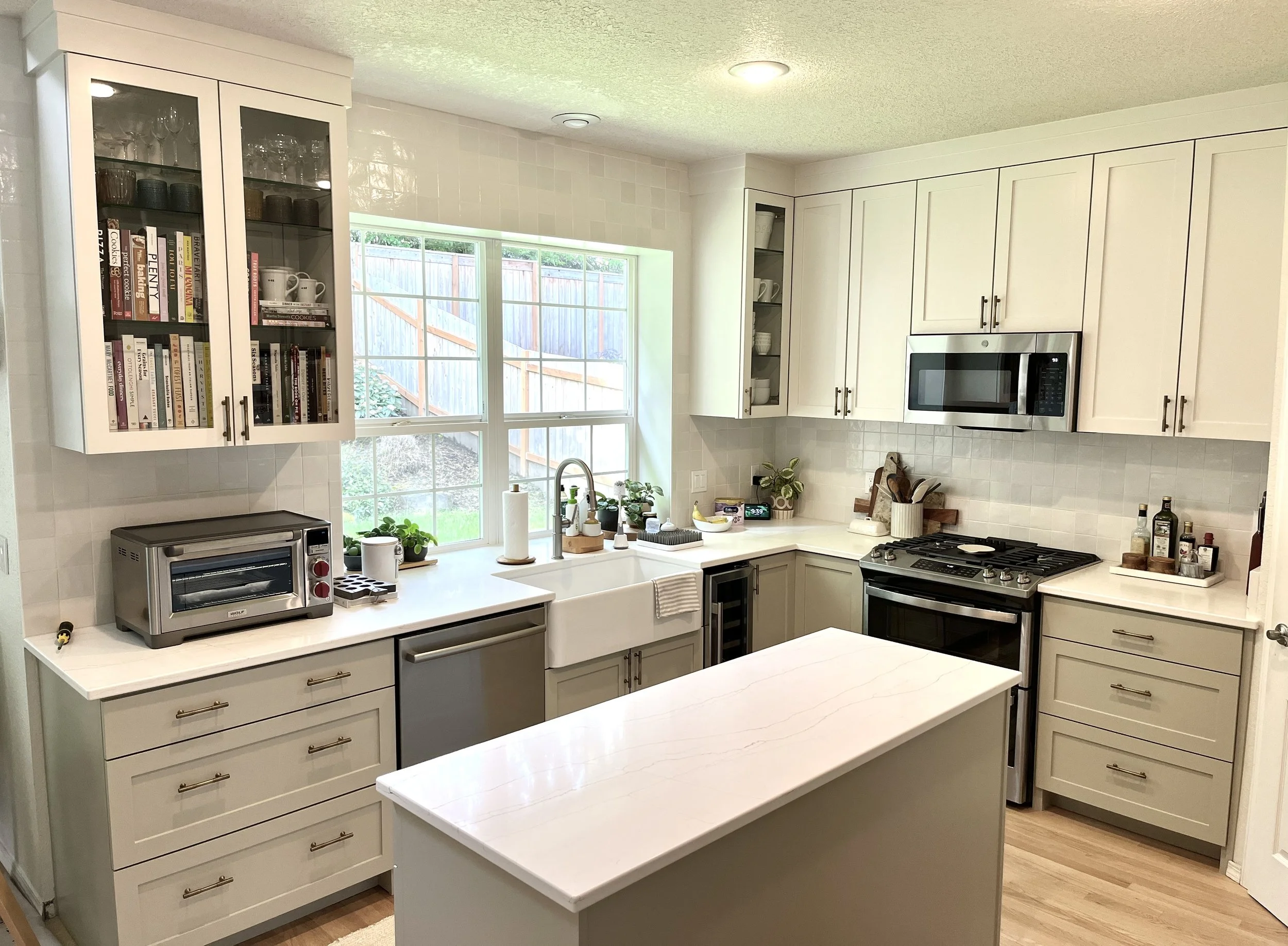 Modern kitchen with white cabinets, a large window, and various appliances including a microwave, toaster oven, and gas stove. Kitchen counter has potted plants, a paper towel roll, and utensils. An island with a white countertop is in the foreground