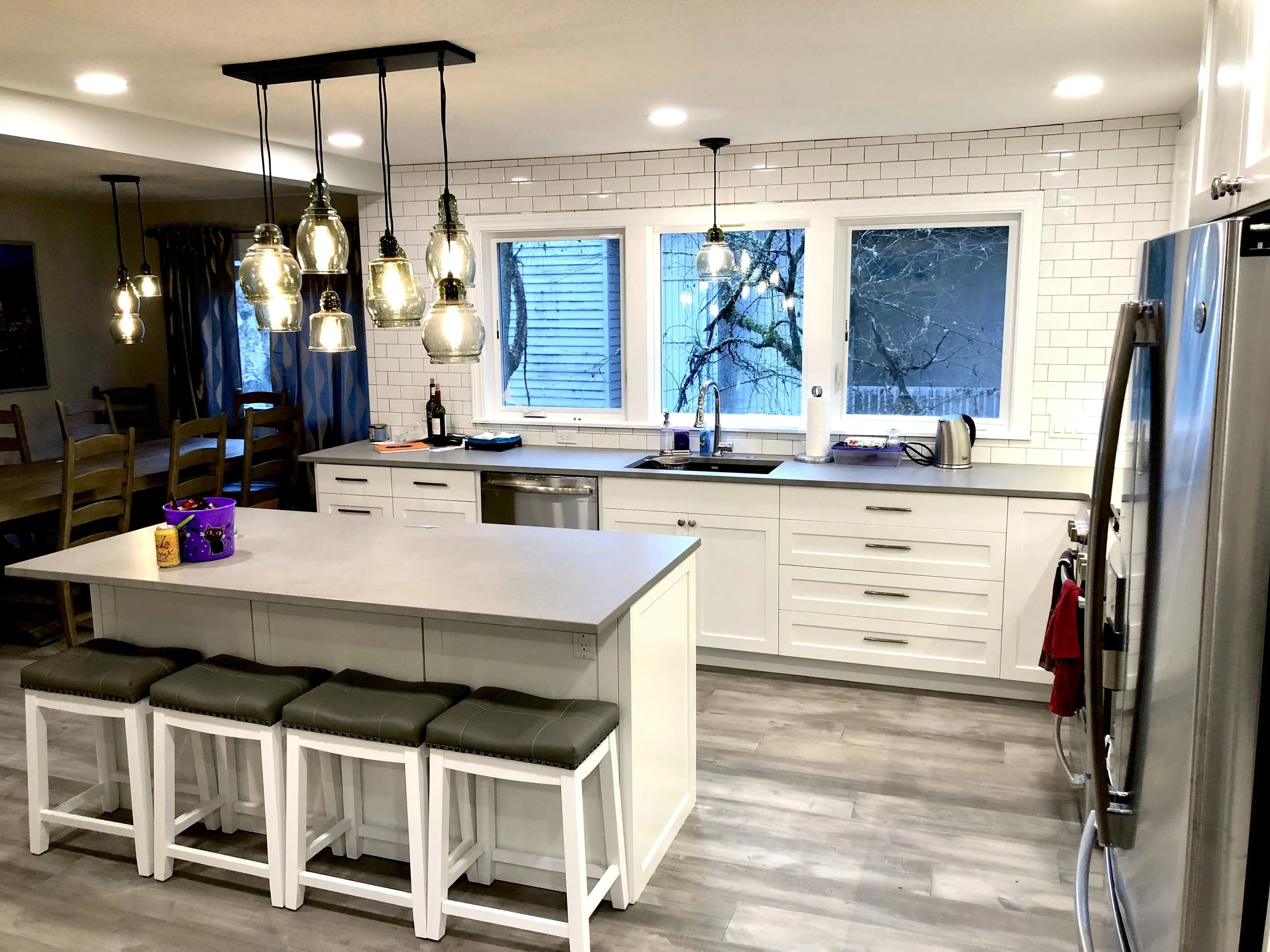 Modern kitchen with white cabinets, a central island with four stools, a window with a view of trees outside, pendant lights hanging over the island, and stainless steel appliances.