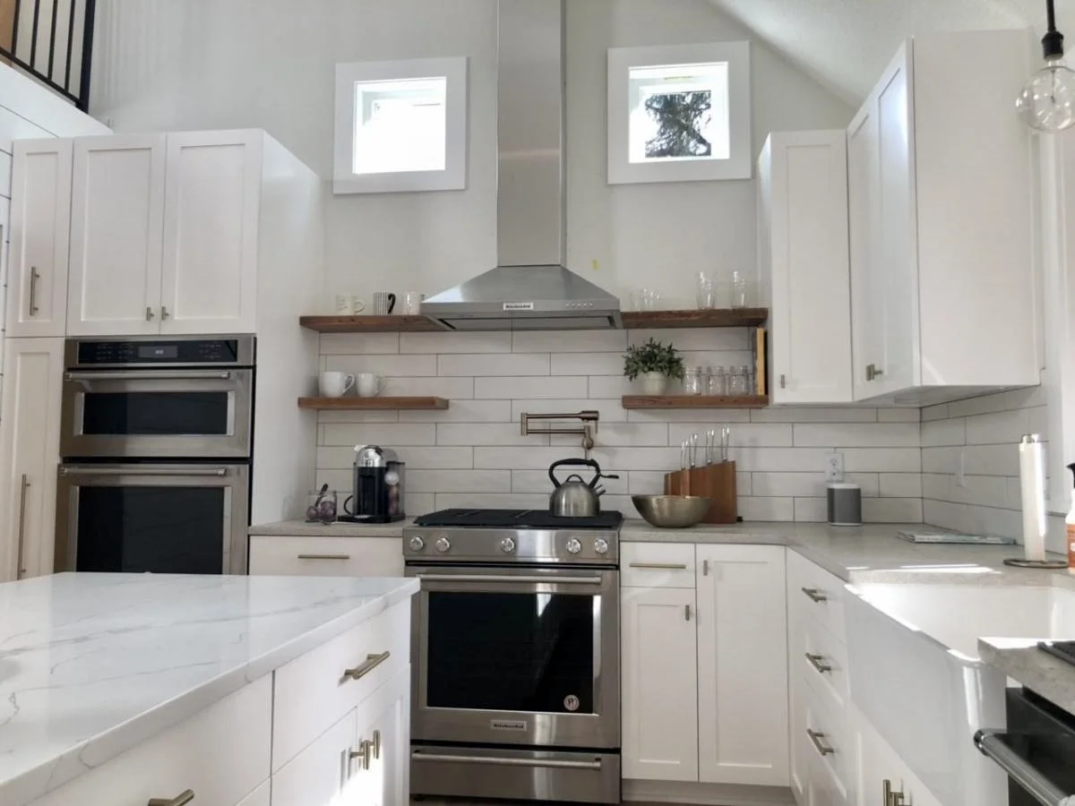 Modern white kitchen with stainless steel oven and stovetop, white cabinets, open wooden shelves with dishes and glassware, and small windows near the ceiling.