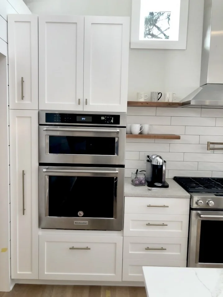 Modern kitchen with white cabinets, stainless steel oven and microwave, countertop with a coffee machine, and wooden shelves holding cups and mugs.