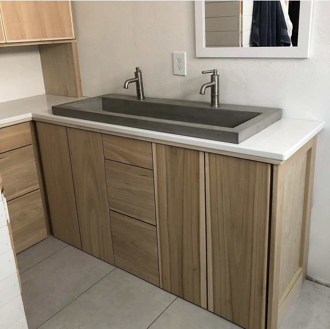 A kitchen sink area with a concrete basin, two stainless steel faucets, wooden cabinets, a white countertop, a mirror, and a wall outlet.
