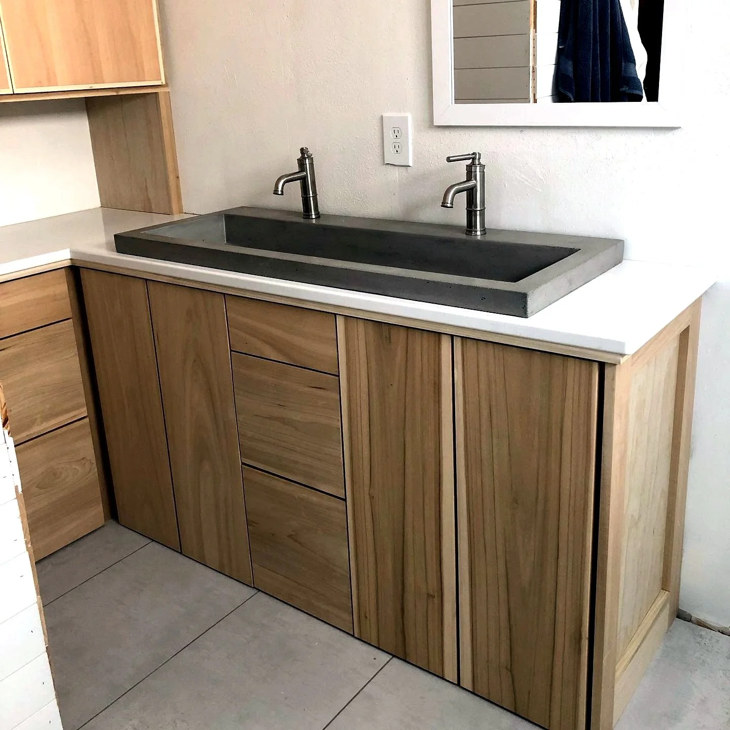 Bathroom vanity with a concrete sink, two modern faucets, wooden cabinetry, a mirror, and a tiled floor.