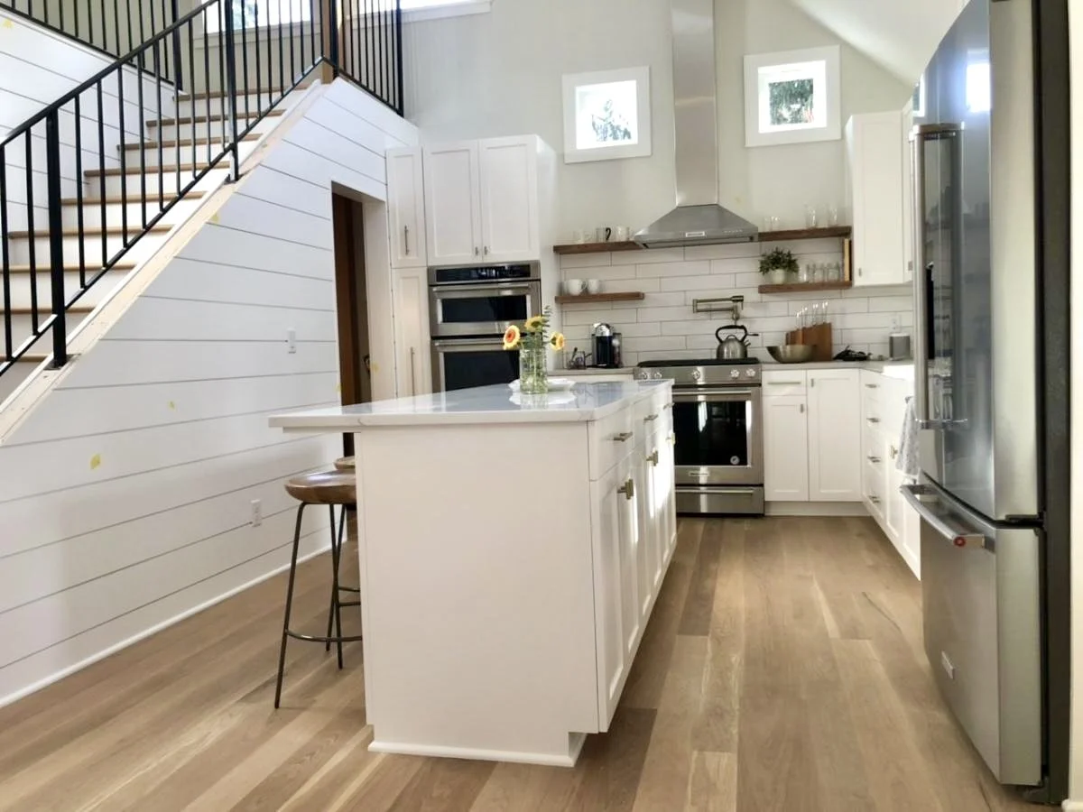 Modern kitchen with white cabinets, stainless steel appliances, a white island with a flower vase, wooden shelves, and a staircase with black railing.