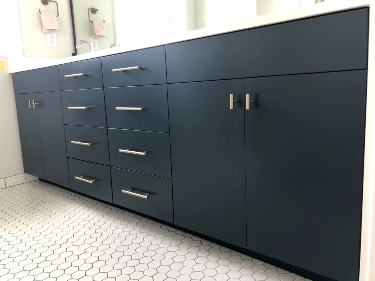 Dark blue bathroom vanity with multiple drawers and cabinet doors, silver handles, white countertop, and hexagonal tile floor.