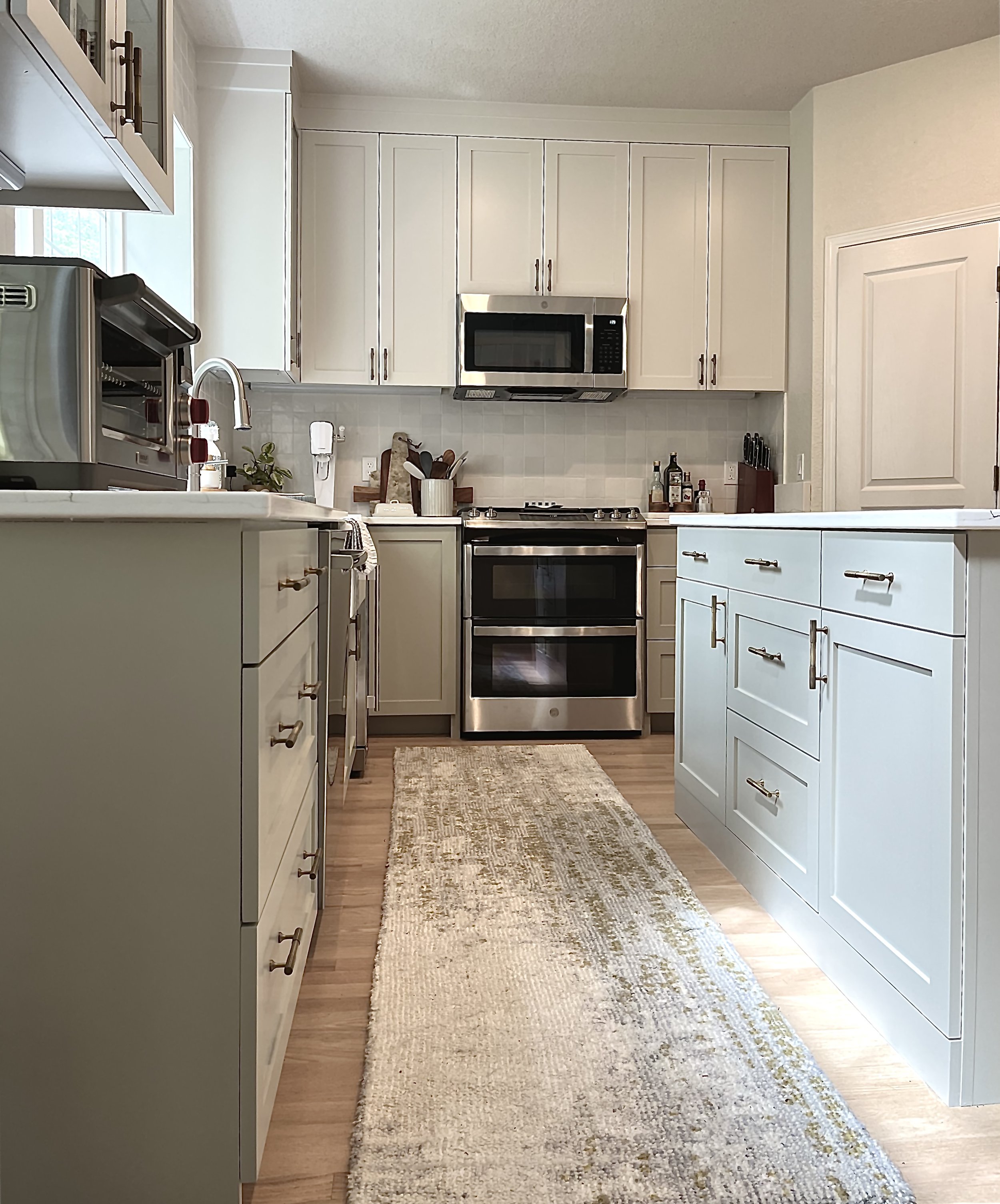 Kitchen with white cabinets, stainless steel stove and microwave, beige backsplash, countertop with small appliances and utensils, beige rug on wooden floor.