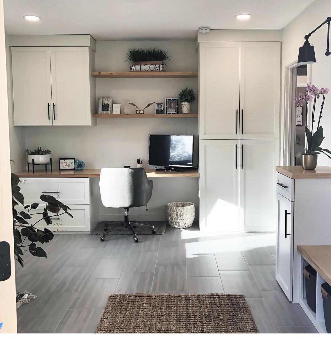Home office setup with white cabinetry, wooden desk, gray swivel chair, small TV, decorative plants, and framed photos on wall shelves.