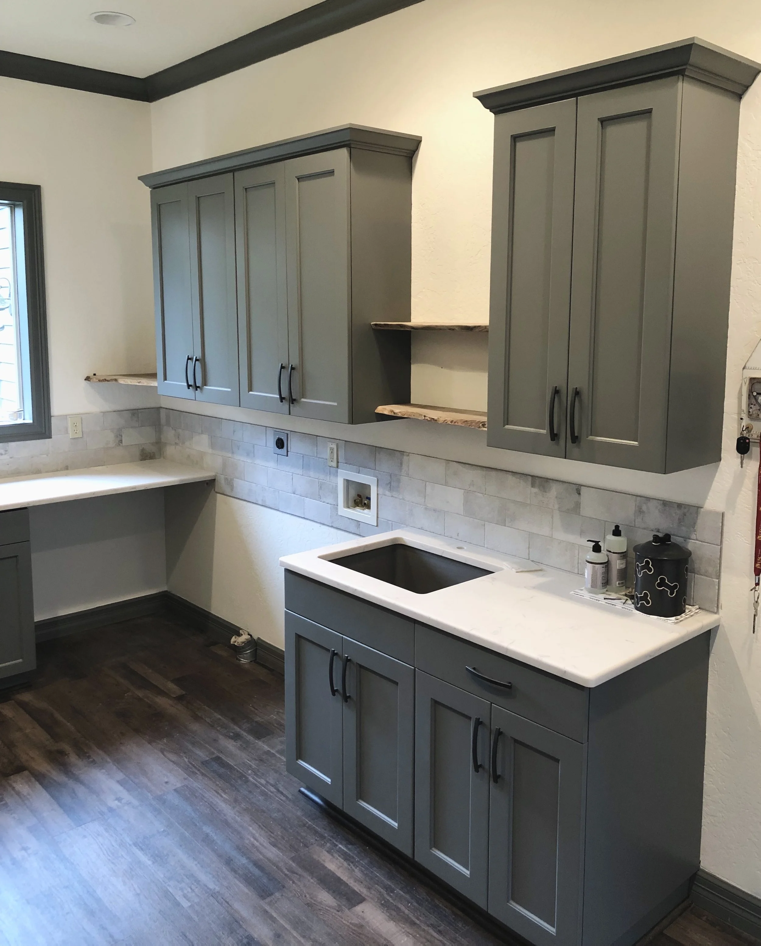 Kitchen with gray cabinets, white marble countertops, a black sink, beige tile backsplash, dark wood flooring, and a window on the left.