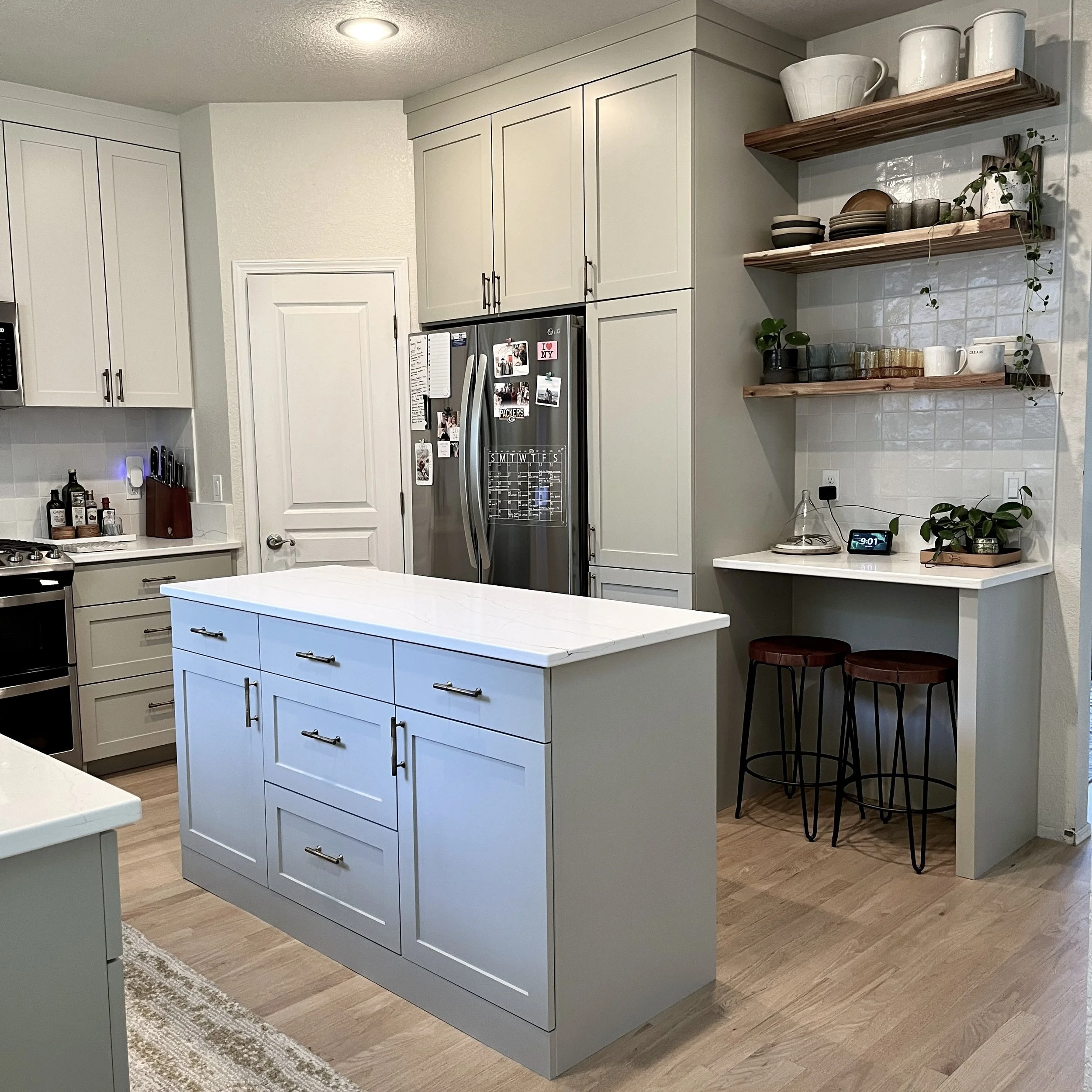 Modern kitchen with light gray cabinets, white island with marble top, stainless steel refrigerator, open wooden shelves with dishes and plants, small workspace with two stools, and wood flooring.